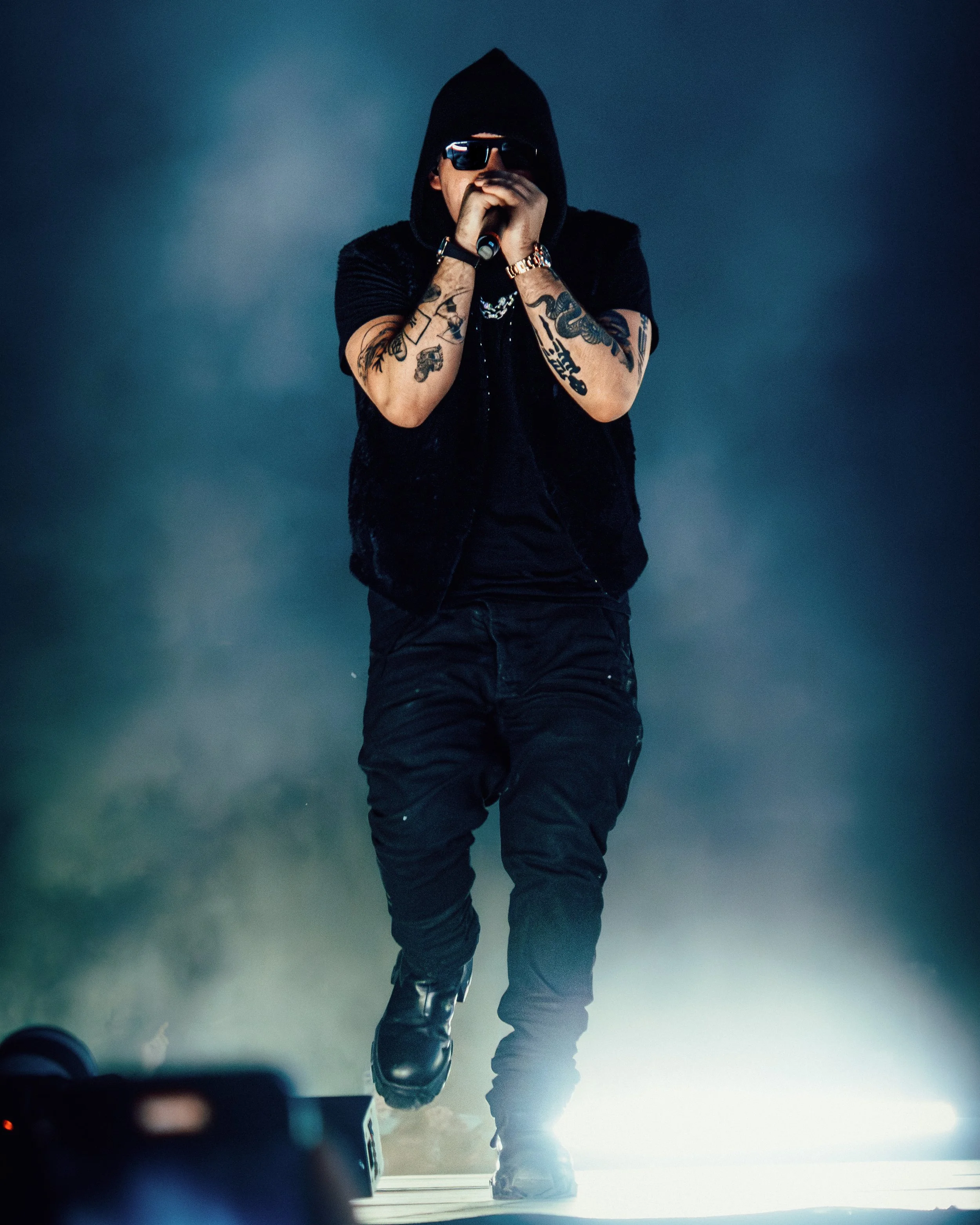 A male performer wearing sunglasses, a black hoodie, and black pants, holding a microphone and performing on stage with a dark background and bright stage lights.