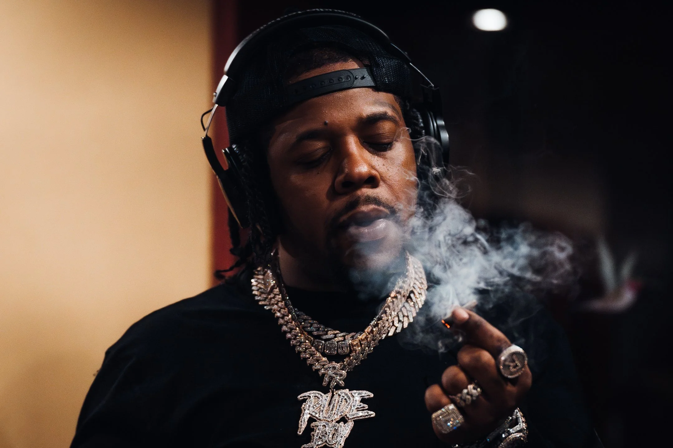 Man wearing headphones and multiple jewelry necklaces and rings, smoking a cigar, with smoke around his face.