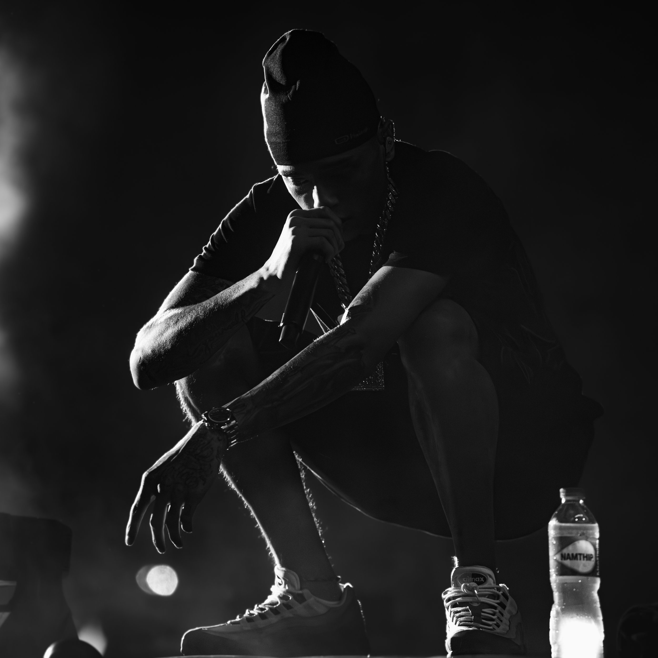 A male rapper crouching on stage, holding a microphone, wearing a beanie, sneakers, and jewelry, with a water bottle next to him, in a dimly lit environment.
