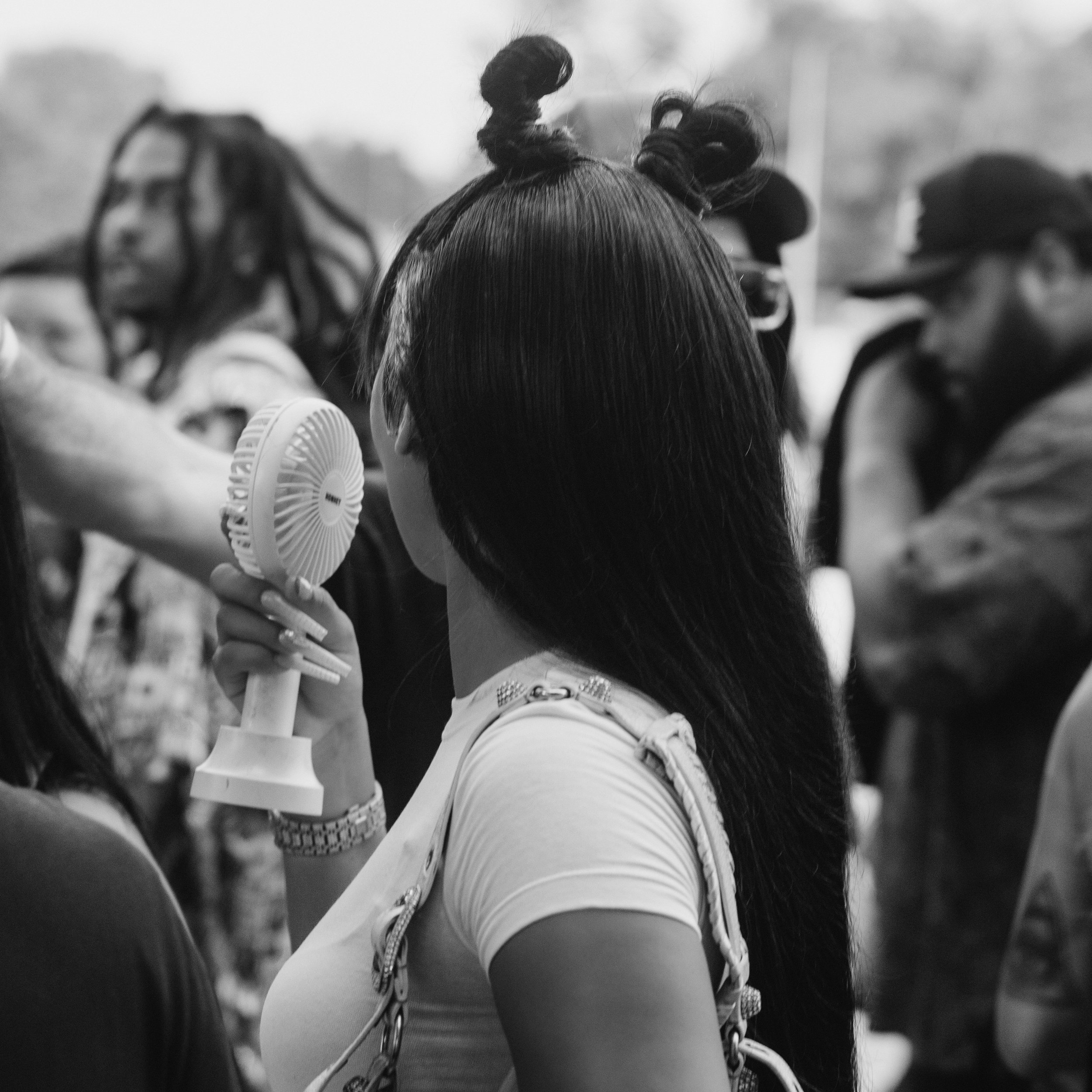 A woman with long dark hair styled in double buns, holding a small fan to her face, surrounded by a crowd in an outdoor setting.