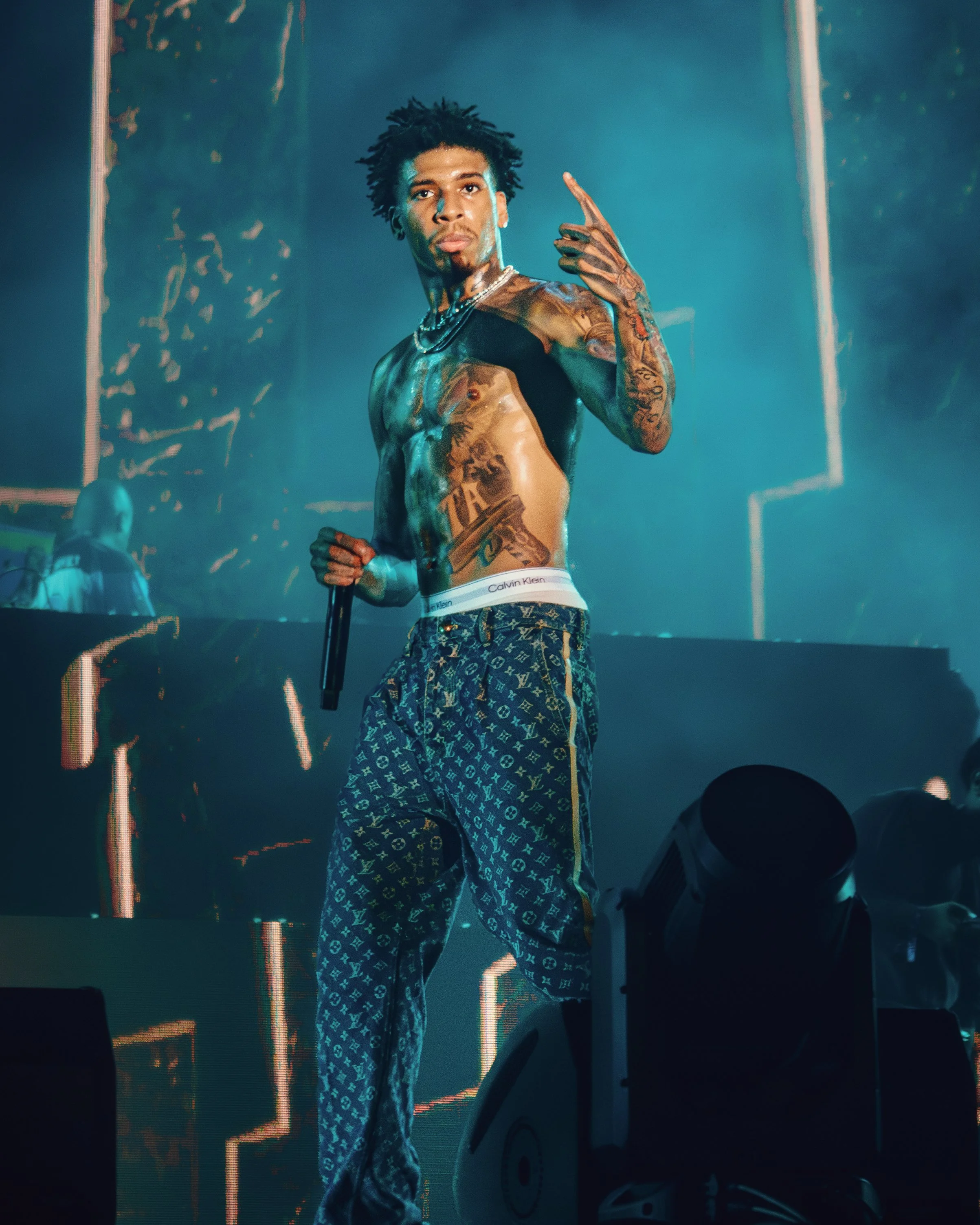 A shirtless male performer with tattoos on his chest and arms holding a microphone on stage during a concert.