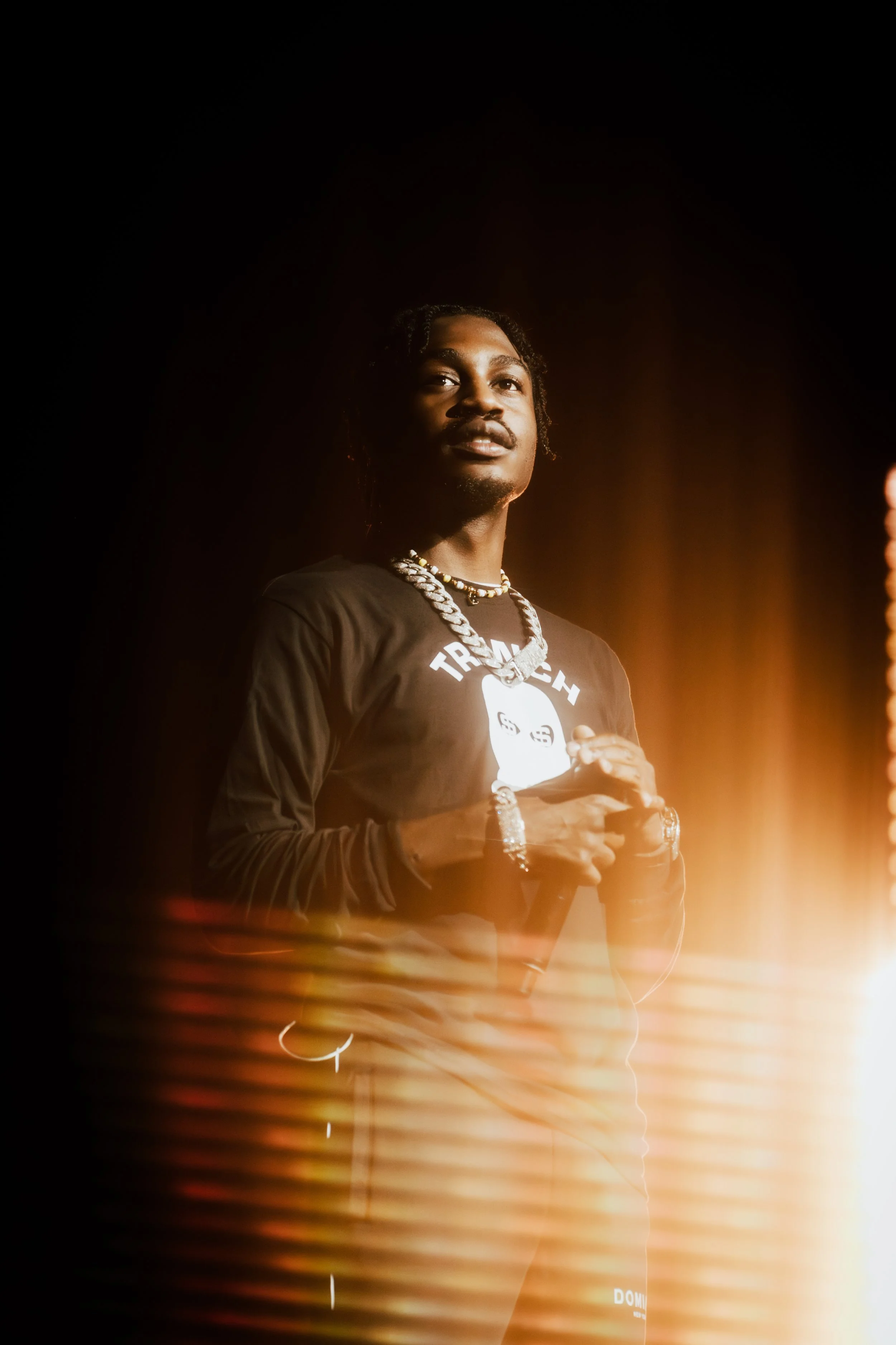 A young man stands against a dark background with warm light illuminating his face and jewelry, wearing a black long-sleeve shirt with a graphic and text on it.