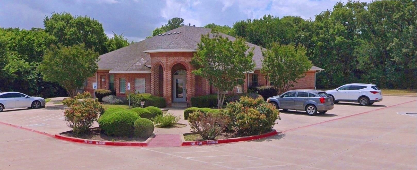A single-story brick building surrounded by a parking lot with several cars parked. The building has a gabled roof and an entrance with an archway. The area is landscaped with bushes and trees, and there are trees in the background.