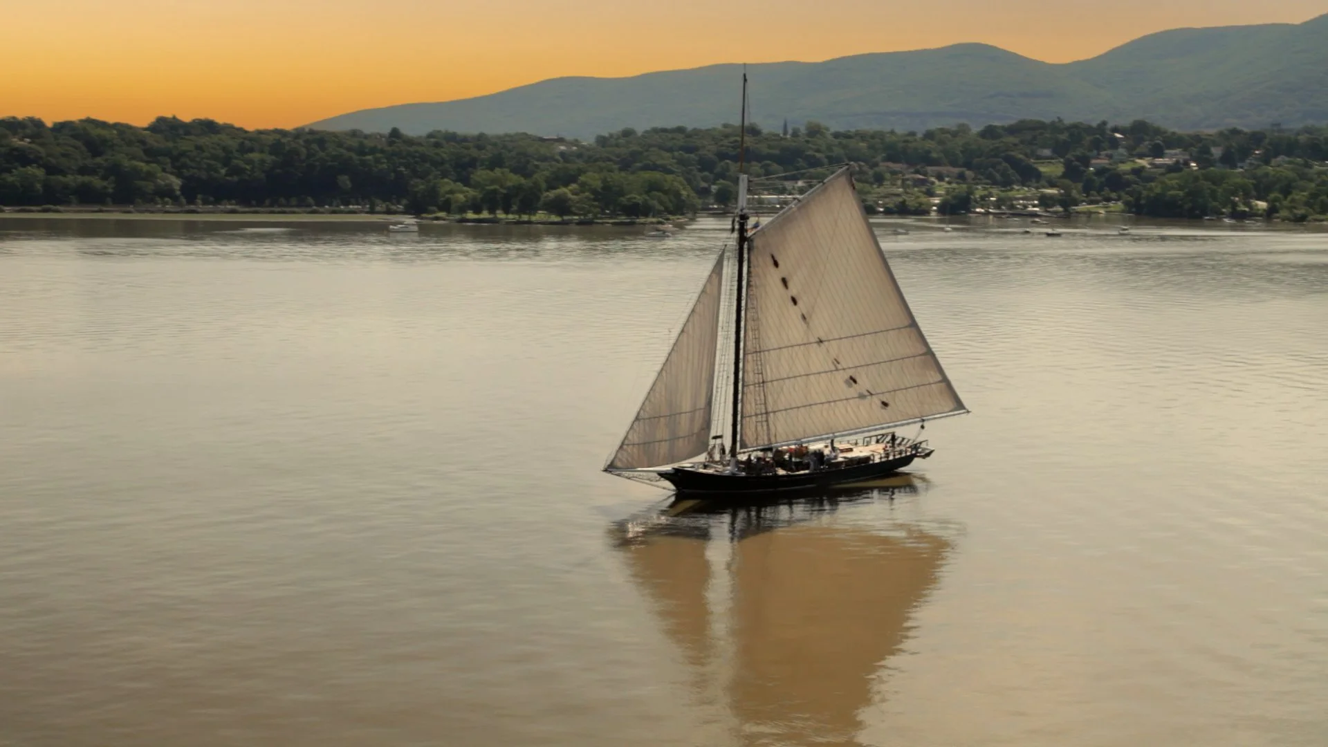 A sailboat with beige sails on a calm river at sunset, mountains and trees in the background.
