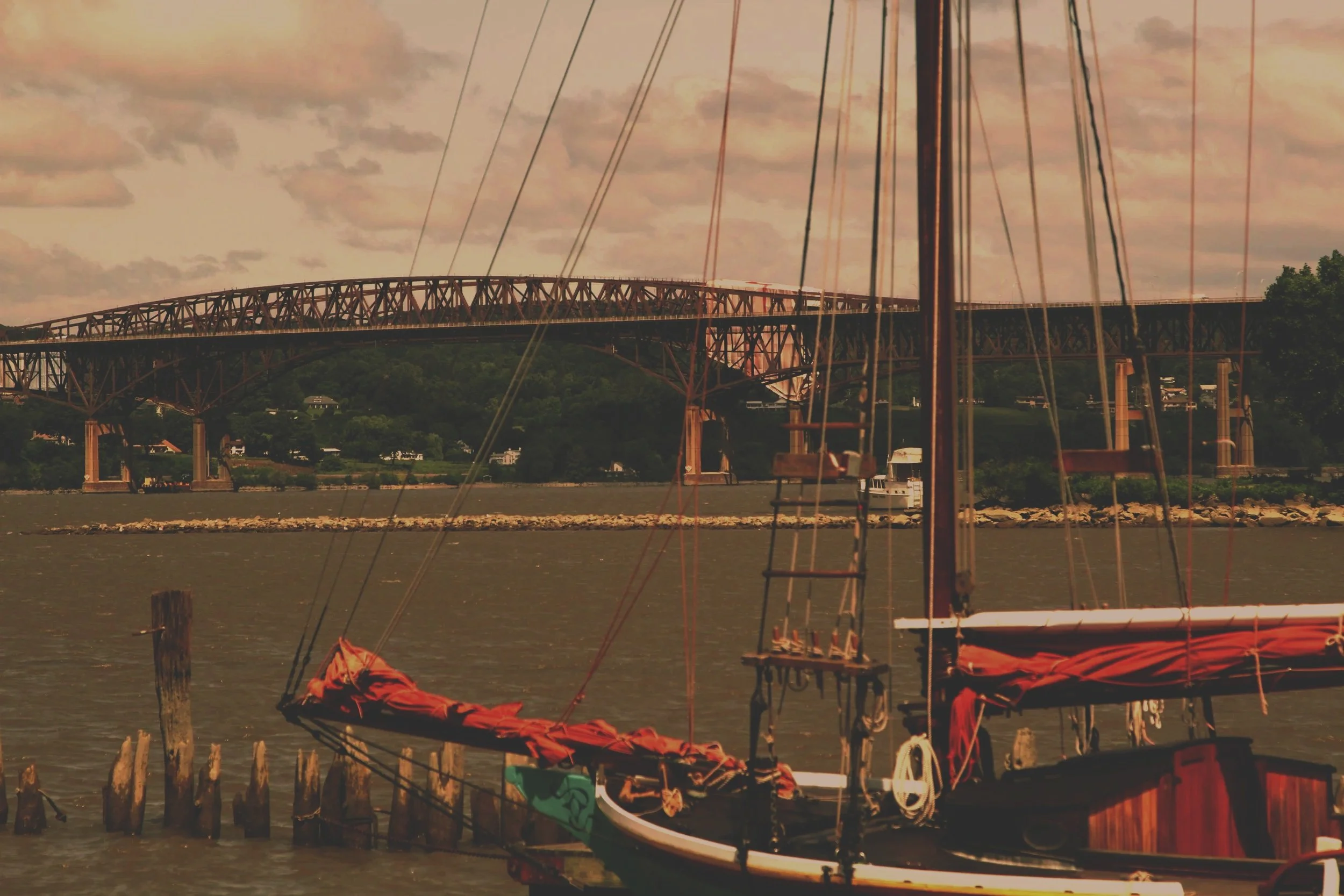 A scene of a bridge over a river with sailboats docked at the shore, power lines in the foreground, and lush green hills in the background under a cloudy sky.