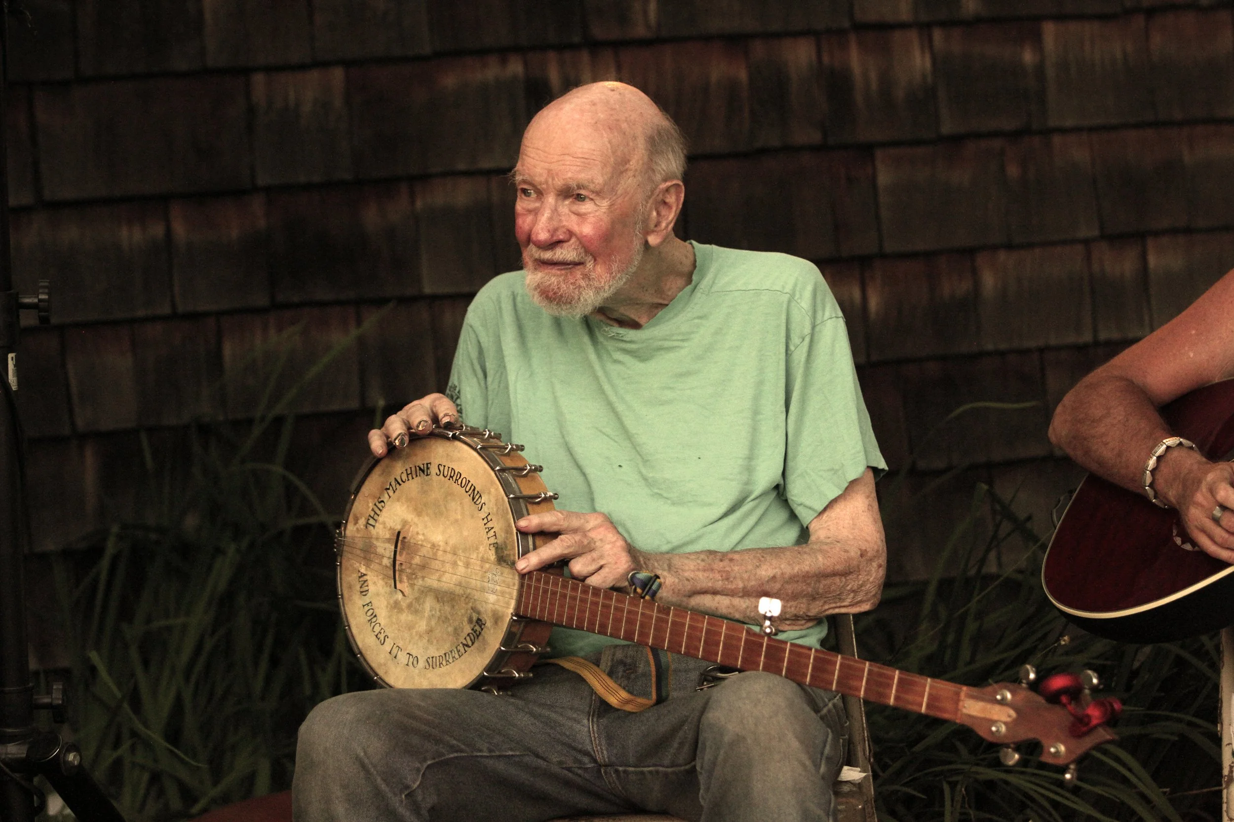 An elderly man with a white beard and receding hairline, wearing a light green t-shirt, sitting and playing a banjo indoors. There is another person partially visible to his right with a guitar. The background consists of a dark wooden shingle wall, 