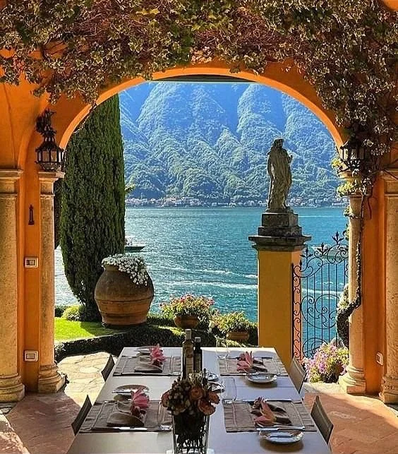 Vista di una terrazza con tavolo apparecchiato, affacciata su un lago e sulle montagne, con statue e fiori decorativi.