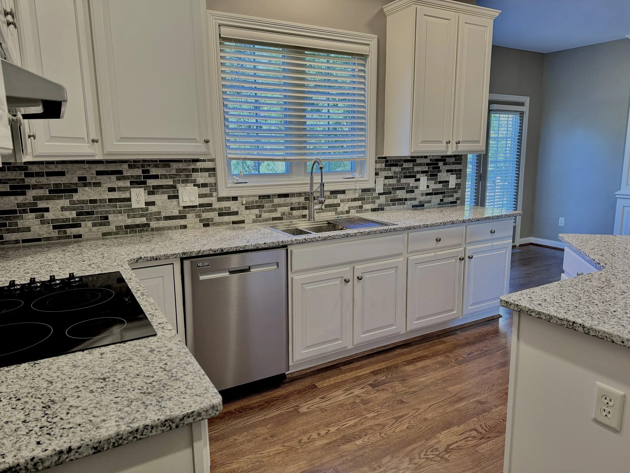 Modern kitchen with granite countertops, white cabinets, stainless steel dishwasher, electric stovetop, and a tiled backsplash. Wood flooring and windows with blinds are visible.