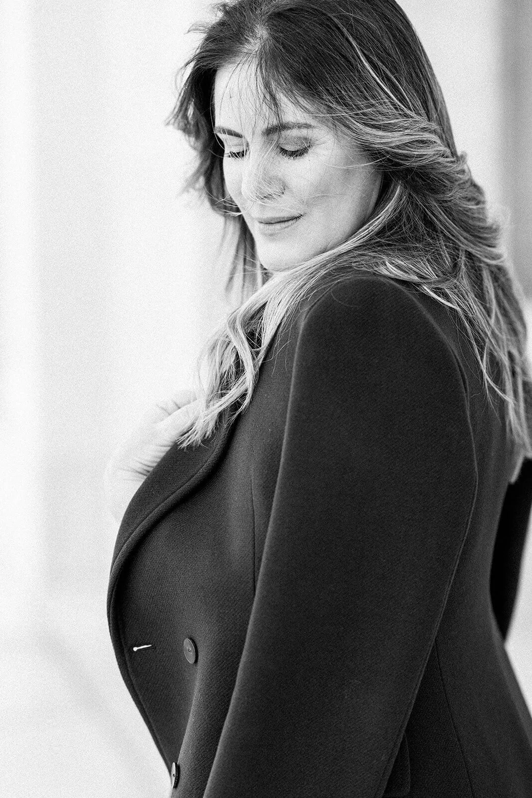 Retrato em preto e branco de uma mulher sorridente com cabelo ondulado, usando blazer preto, de olhos fechados e expressão serena.