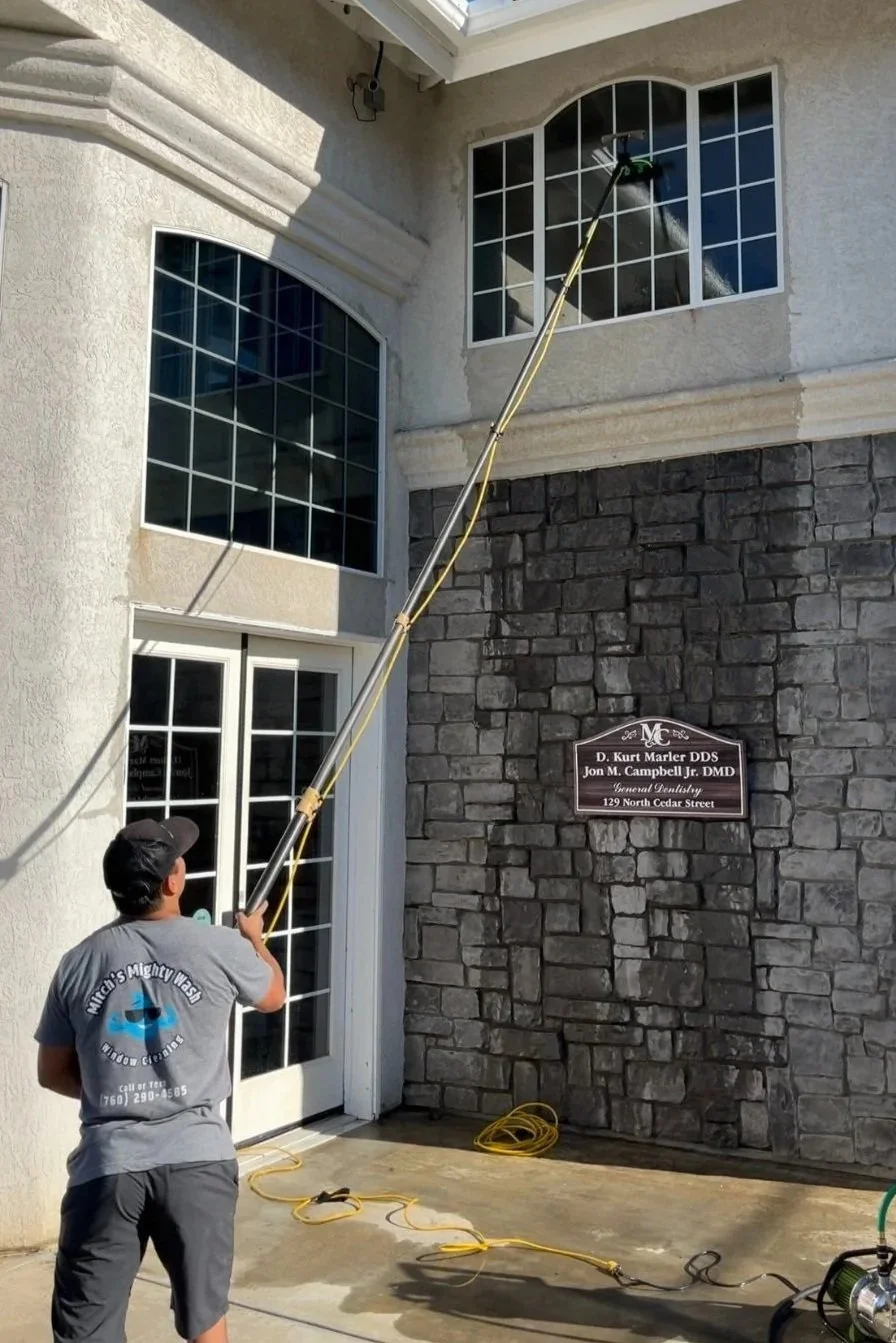 A man cleaning windows on the second story of a building using a long-handled cleaning tool. The building features large windows and a stone and stucco exterior. Cords and cleaning equipment are on the ground.