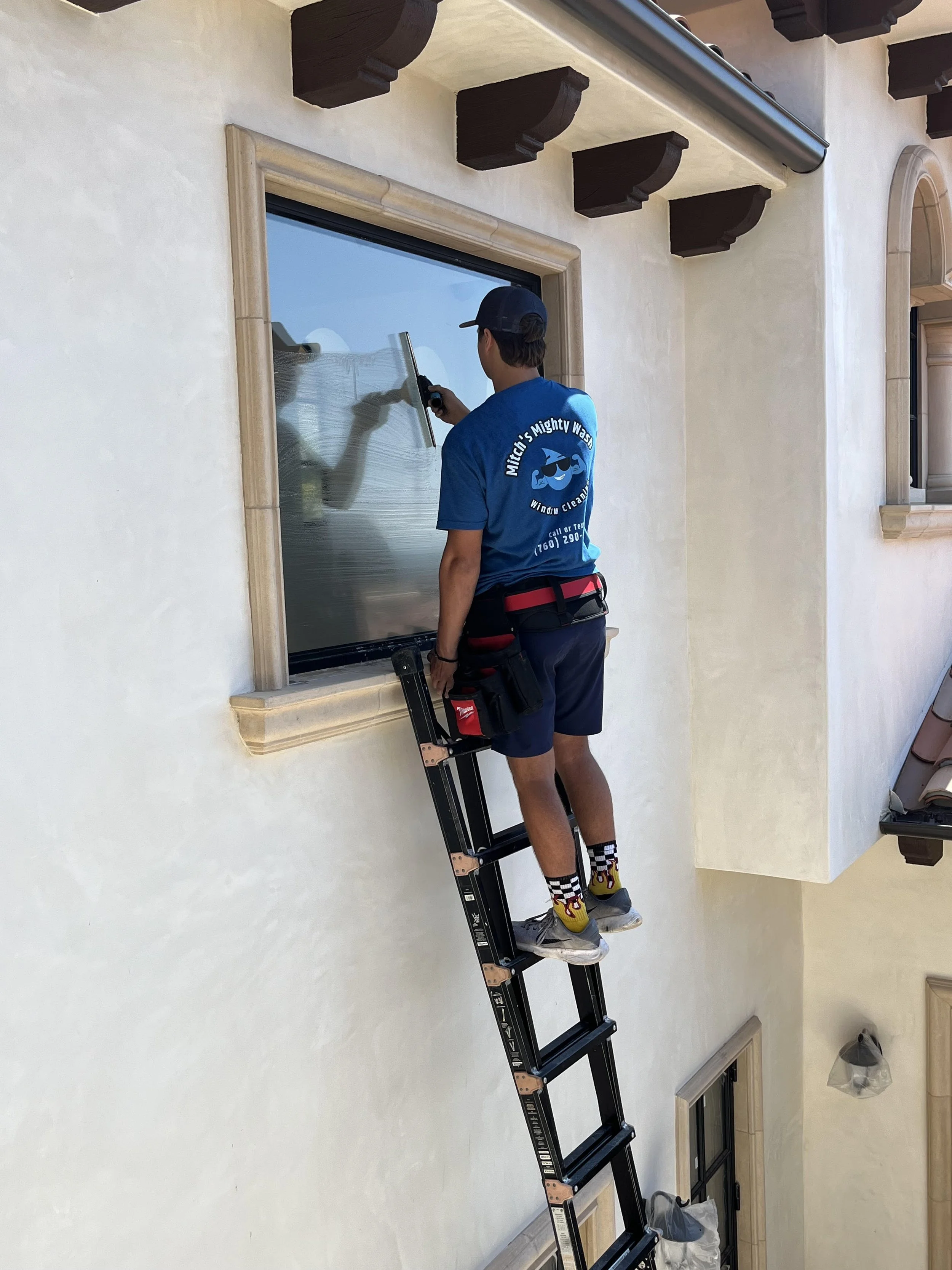 A worker cleaning a window from a ladder on the exterior of a house.