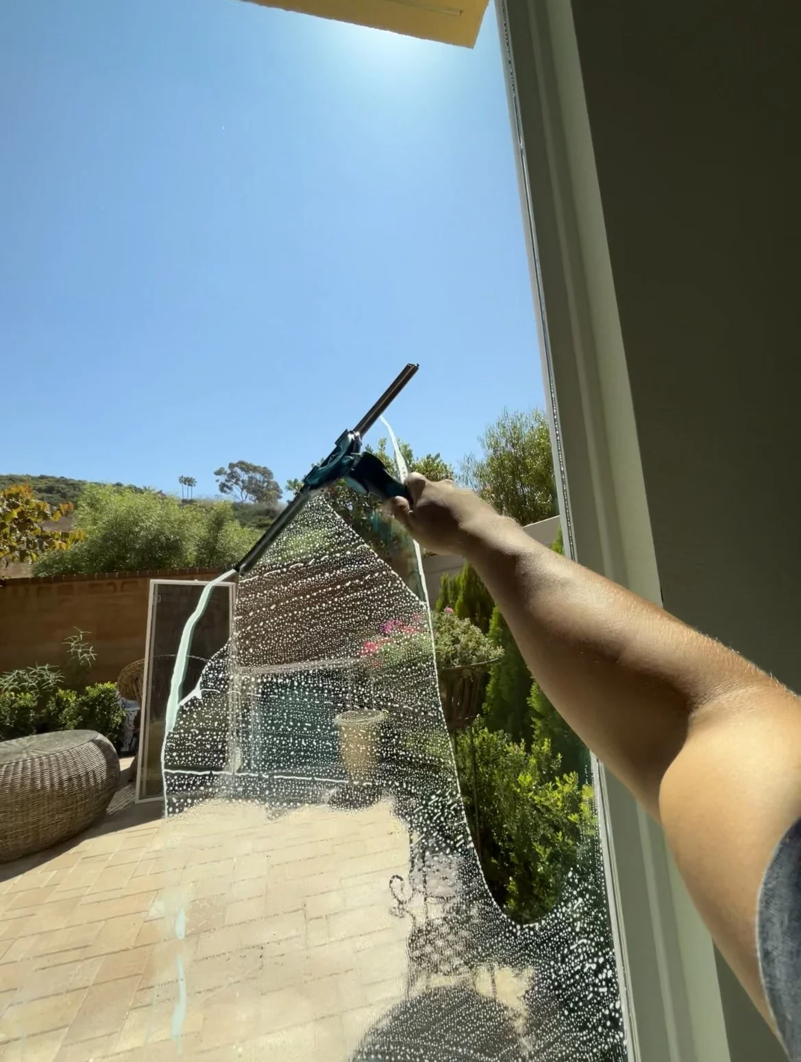 Someone cleaning a glass window with a squeegee, outdoor patio with plants, blue sky, and trees in the background.
