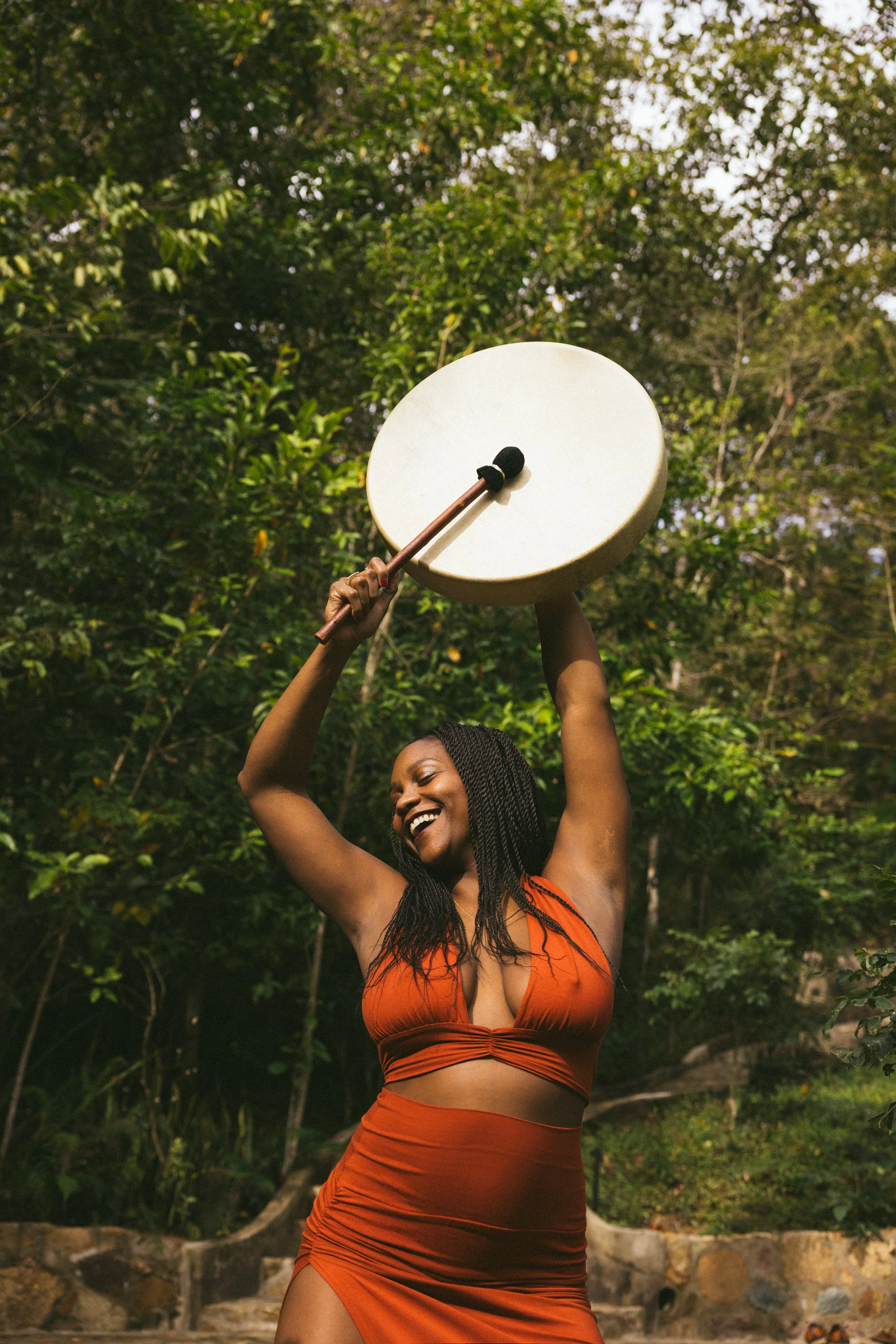 A woman wearing an orange dress holding a round white drum above her head outdoors surrounded by trees.