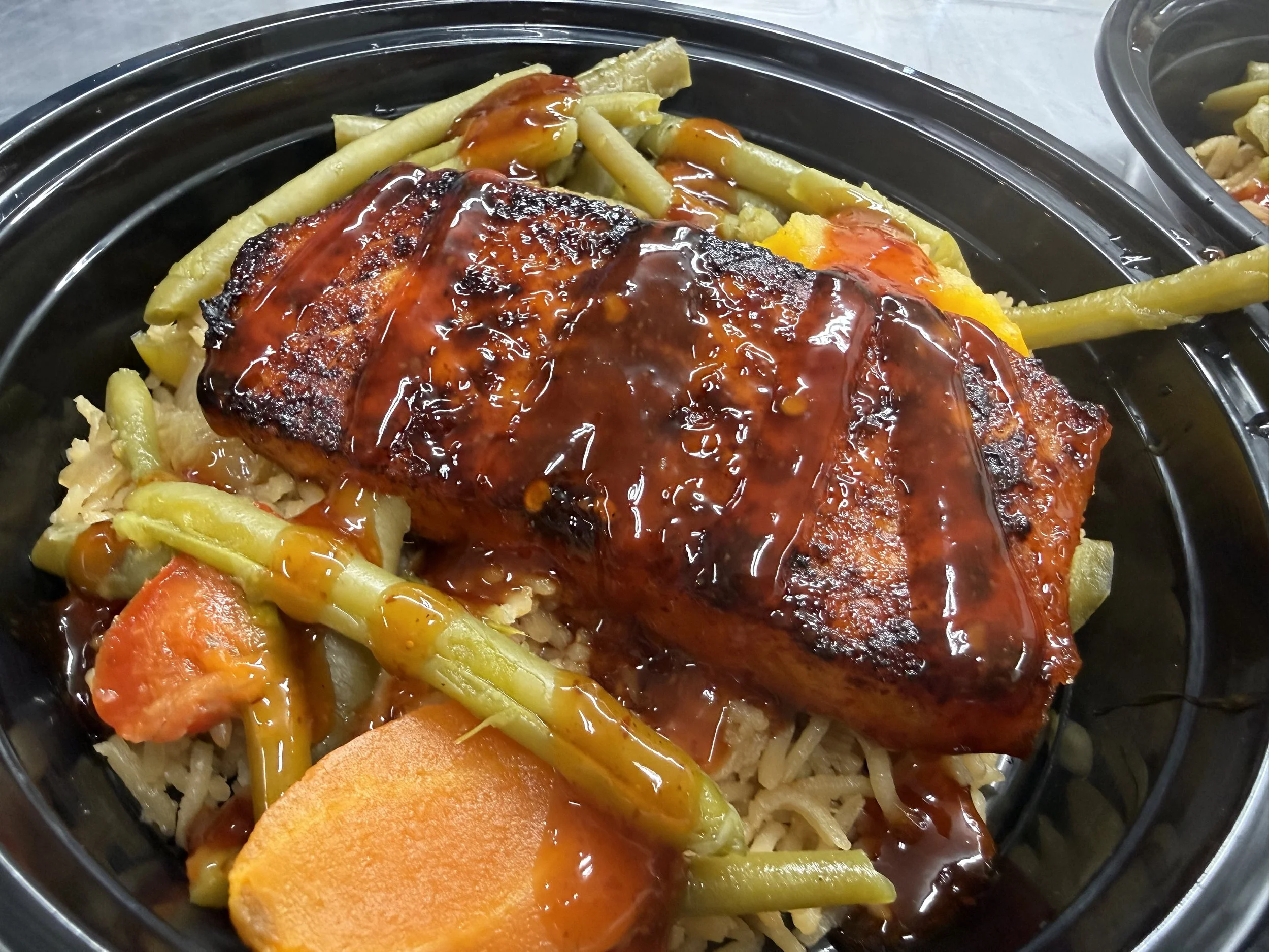 A black plastic takeout container filled with rice, mixed vegetables, a grilled piece of meat with barbecue sauce, and a slice of cooked sweet potato.