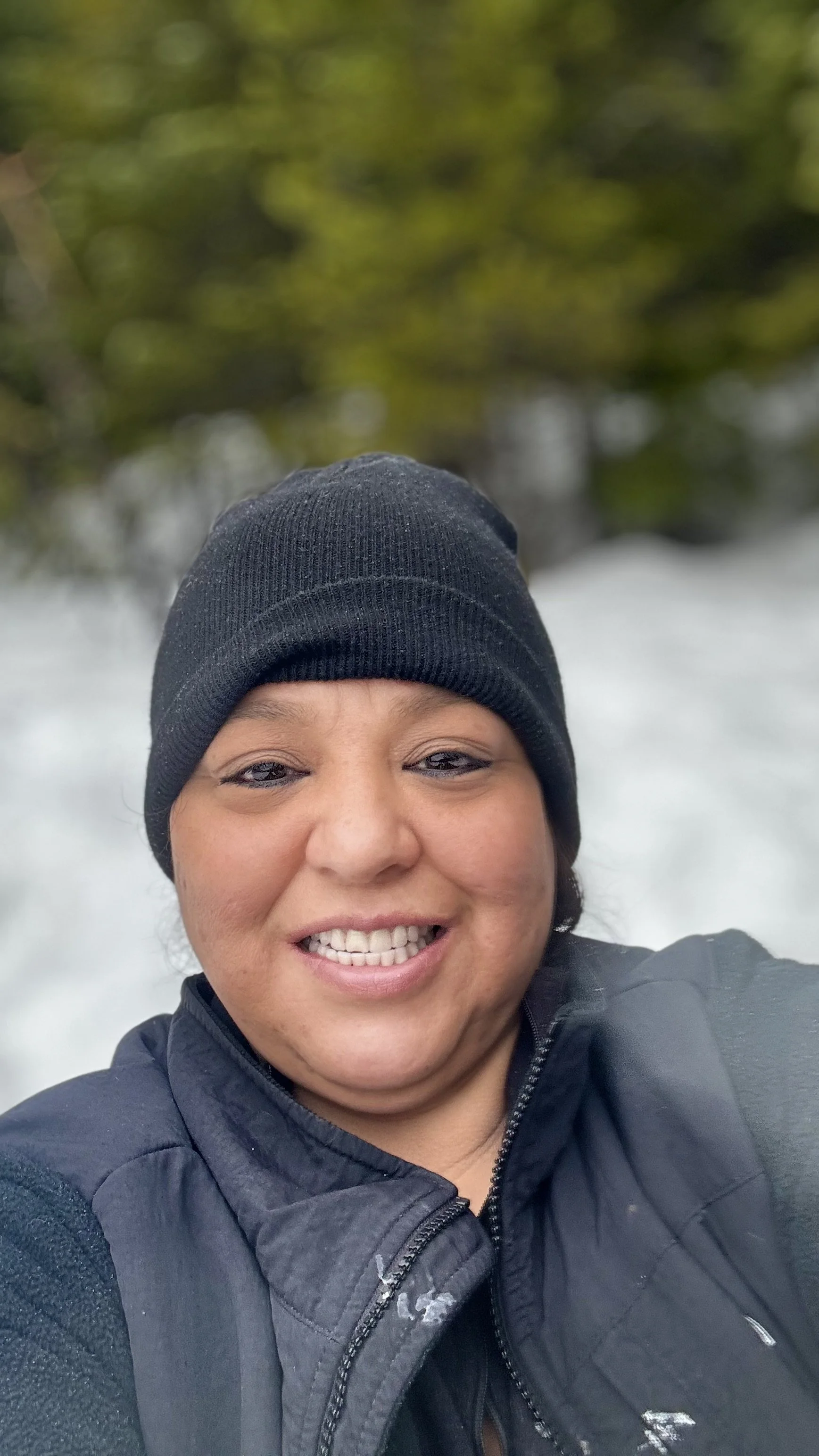 A woman taking a selfie outdoors in a natural setting, wearing a black beanie and a black zip-up jacket, smiling with trees and water in the background.