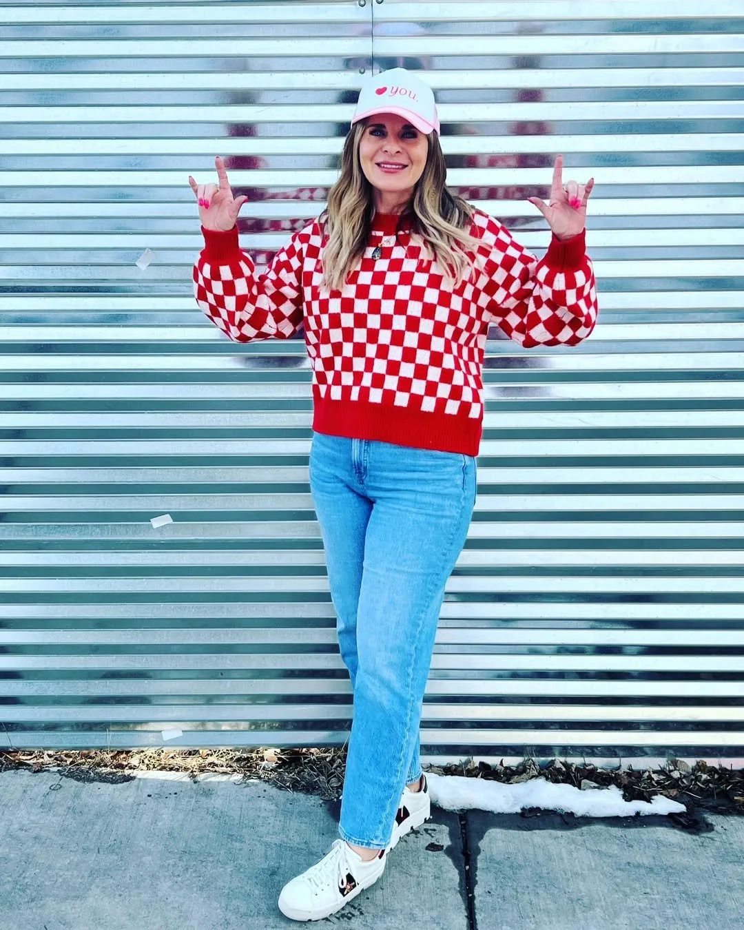Woman standing in front of a metal wall, wearing a red and white checkered sweater, light blue jeans, white sneakers, and a light pink cap that says 'you' with a red heart. She is smiling and making a rock-and-roll hand gesture.