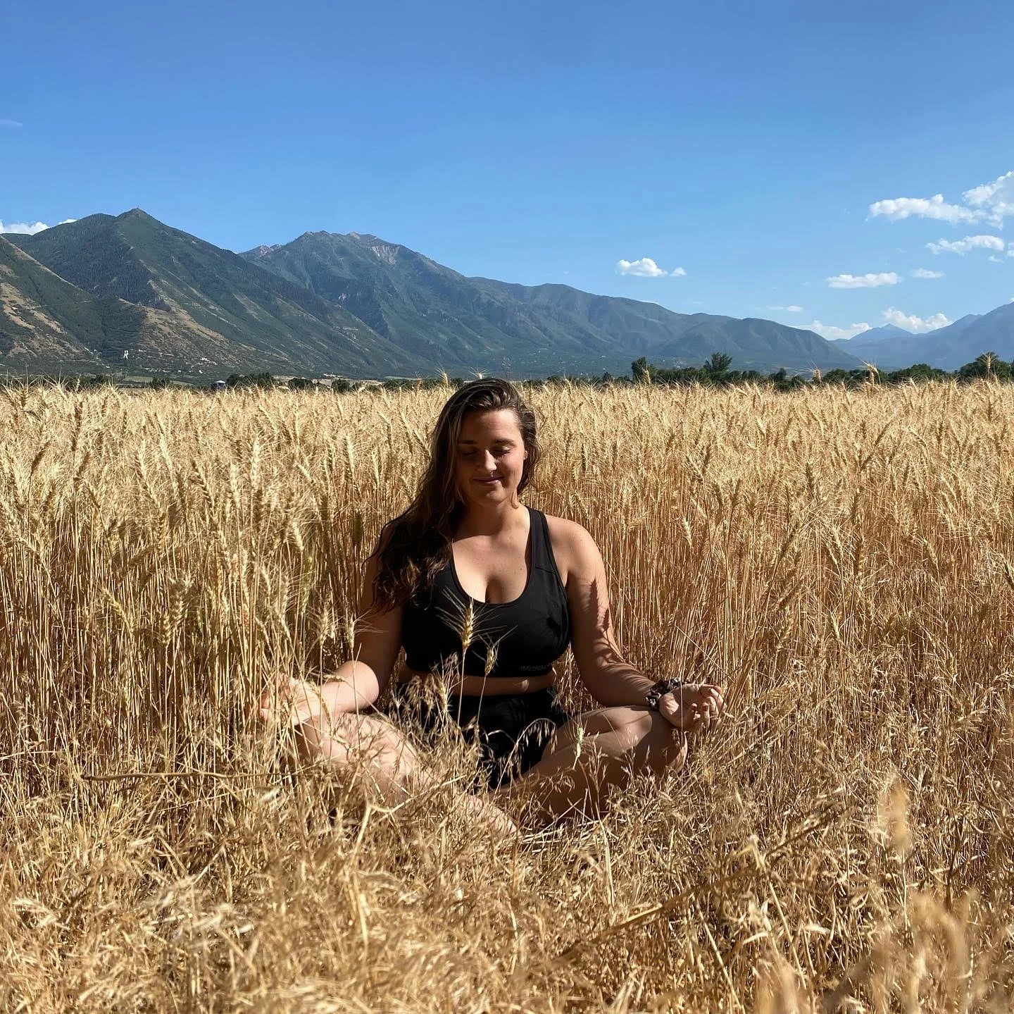 A woman sitting cross-legged in a wheat field with mountains in the background under a clear blue sky.