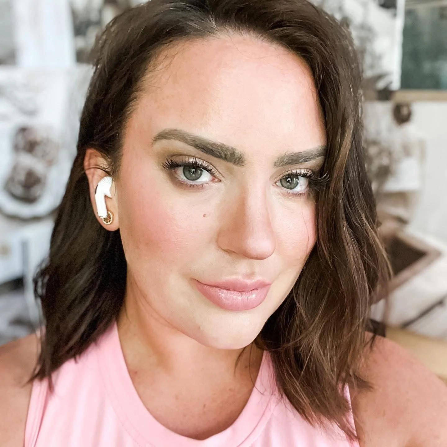 A close-up of a woman with wavy dark brown hair, light makeup, and earrings, wearing a pink top. She has grey eyes and a slight smile.