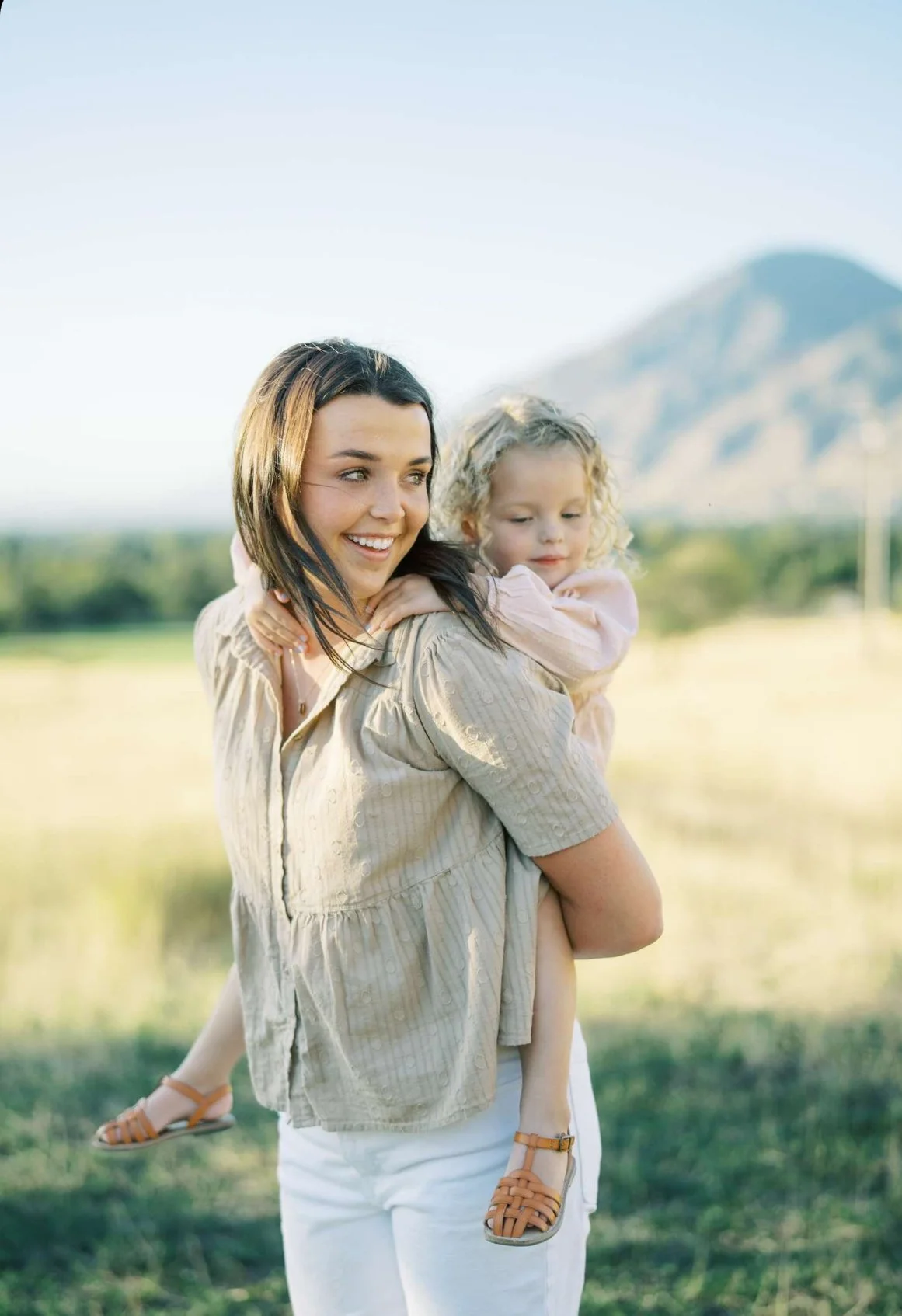 A smiling woman carrying a young girl on her back in a grassy outdoor area with mountains in the background.