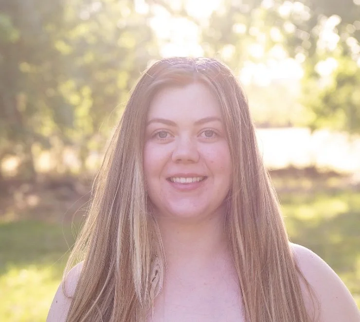 Young woman with long blonde hair smiling outdoors with sunlight and trees in the background.