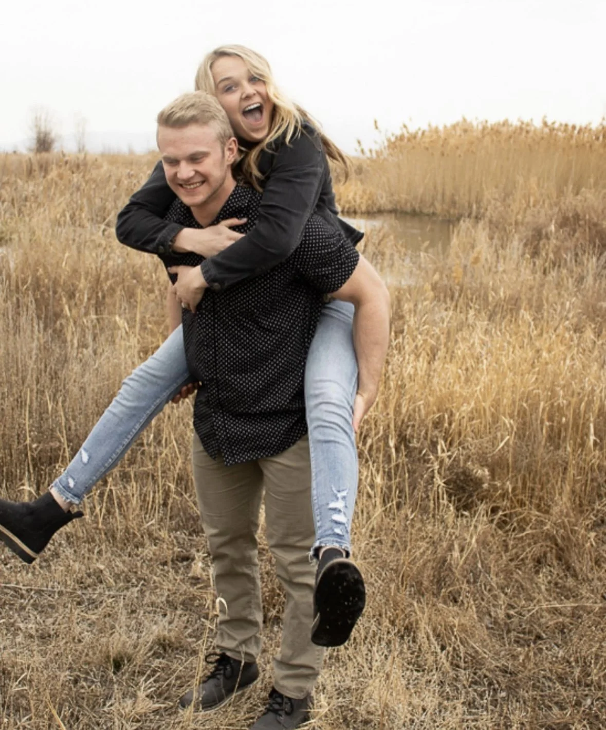 A young couple enjoying a fun moment outdoors in a grassy field, with the woman on the man's back, both smiling and laughing.