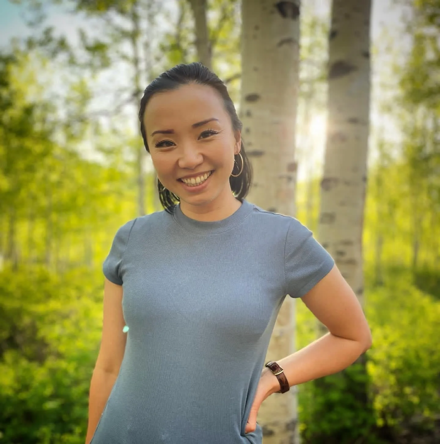 A woman smiling in a forest with green trees and sunlight in the background.