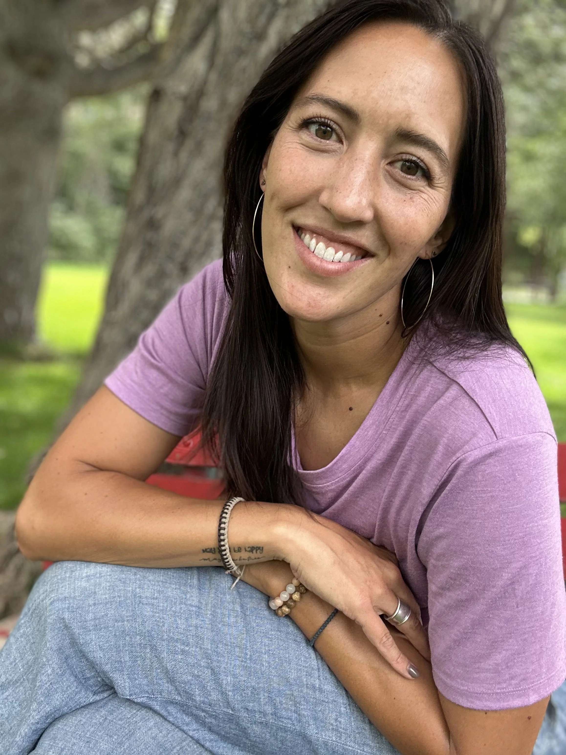 A woman smiling outdoors near a large tree, wearing a pink T-shirt, hoop earrings, and jewelry, with green grass and trees in the background.
