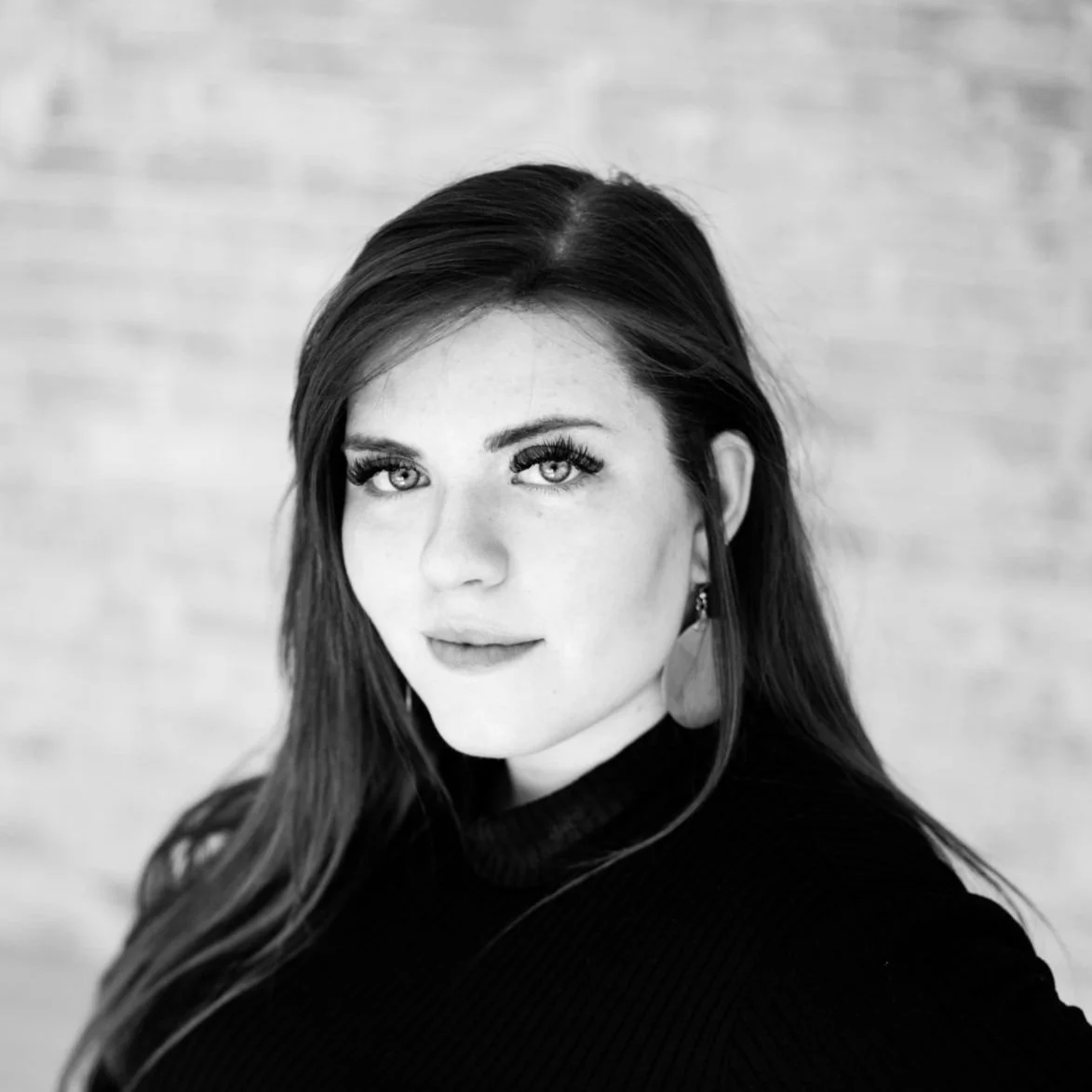 Black and white portrait of a woman with long hair, wearing large earrings and a black turtleneck, with a blurred brick wall background.