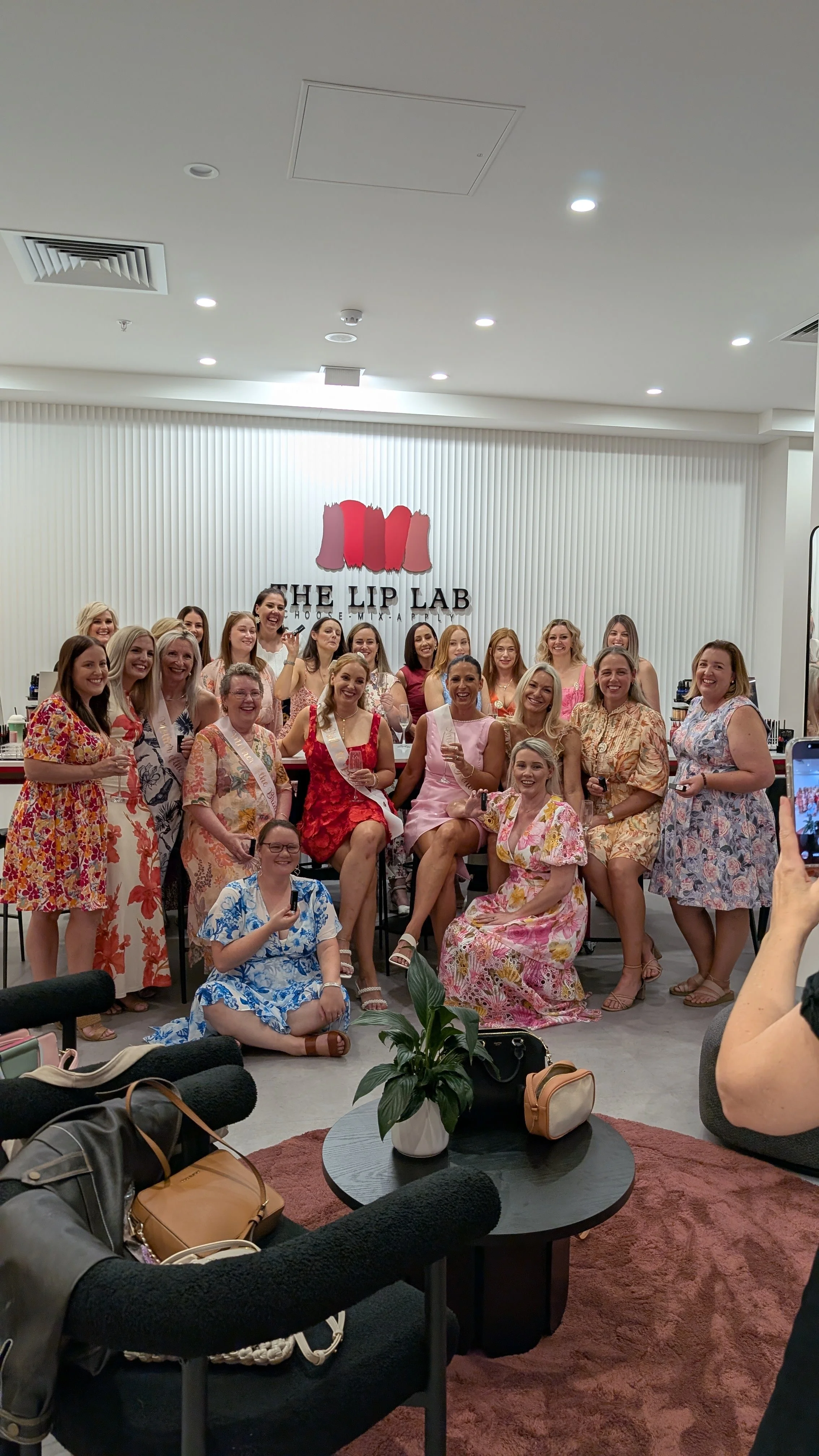 Group of women in colorful dresses celebrating at The Lip Lab, some holding drinks, smiling, and posing for a photo in a modern room with a white wall and logo.