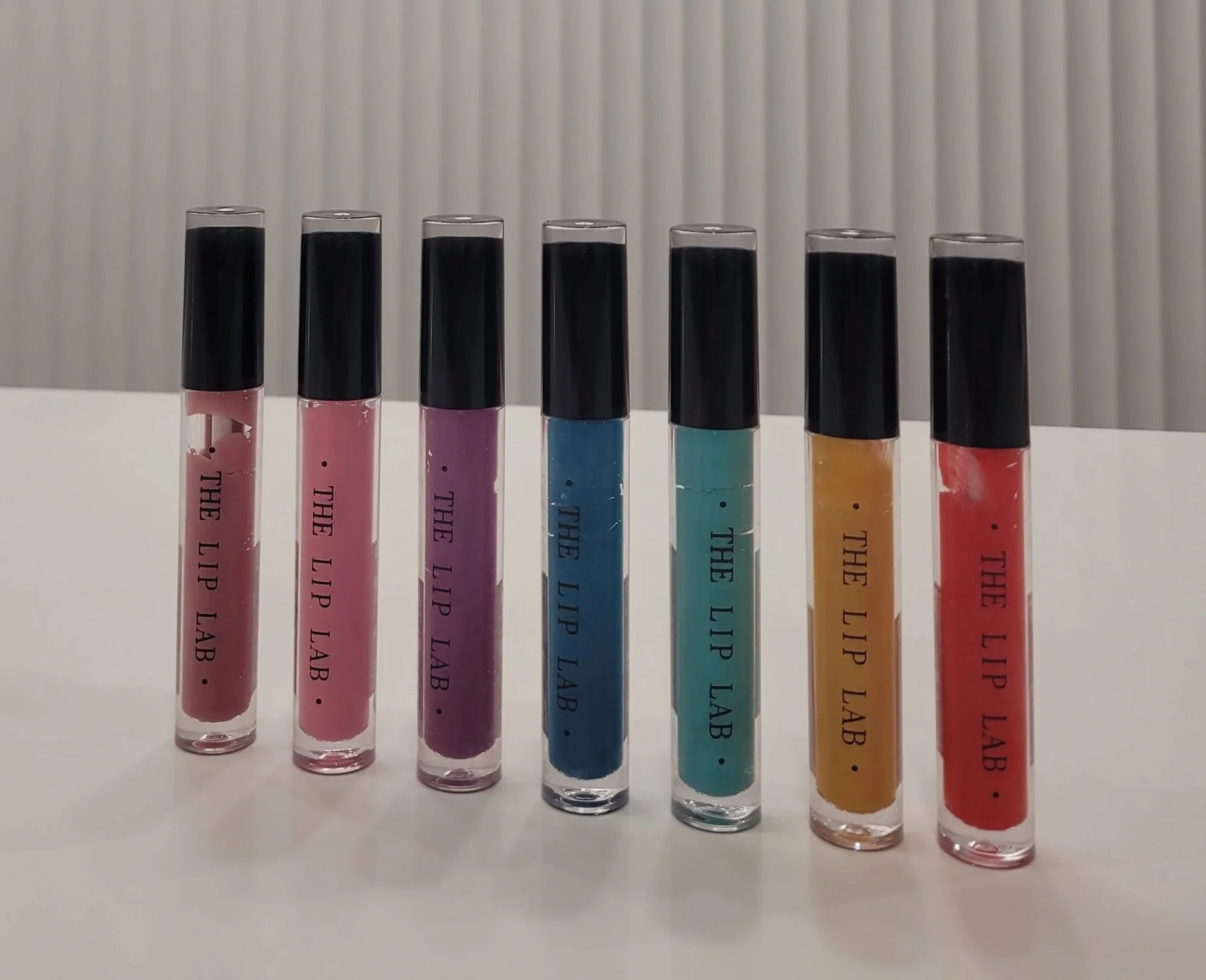 Six colourful lip gloss tubes with black caps, arranged in a line on a white surface with a vertically striped gray background.