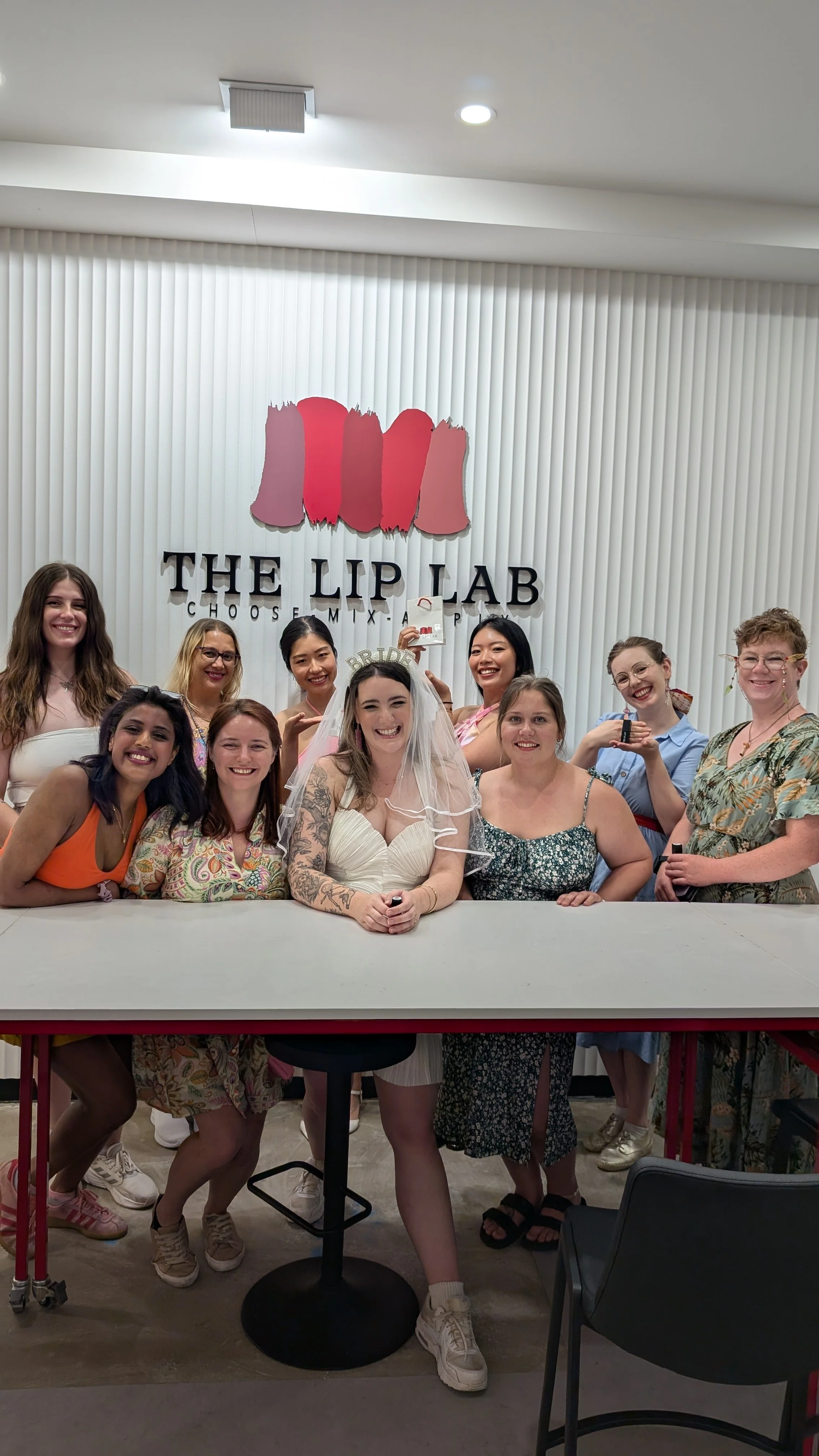 Group of women, including a bride in a wedding dress wearing a crown that says 'BRIDE', posing for a photo at The Lip Lab, with a backdrop featuring a large logo with three lipstick shapes and the words 'THE LIP LAB CHOOSE. MIX. APPLY'.