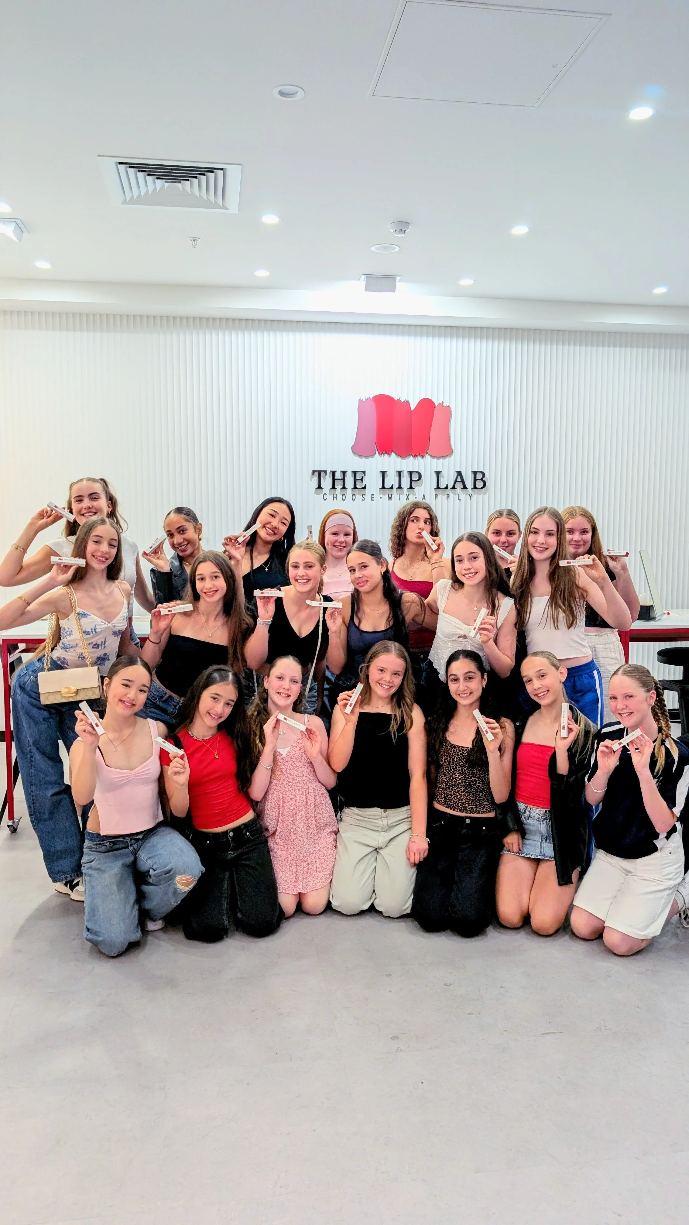 A group of young girls posing for a photo inside a store or salon called THE LIP LAB, with some holding lip gloss or lipstick products.