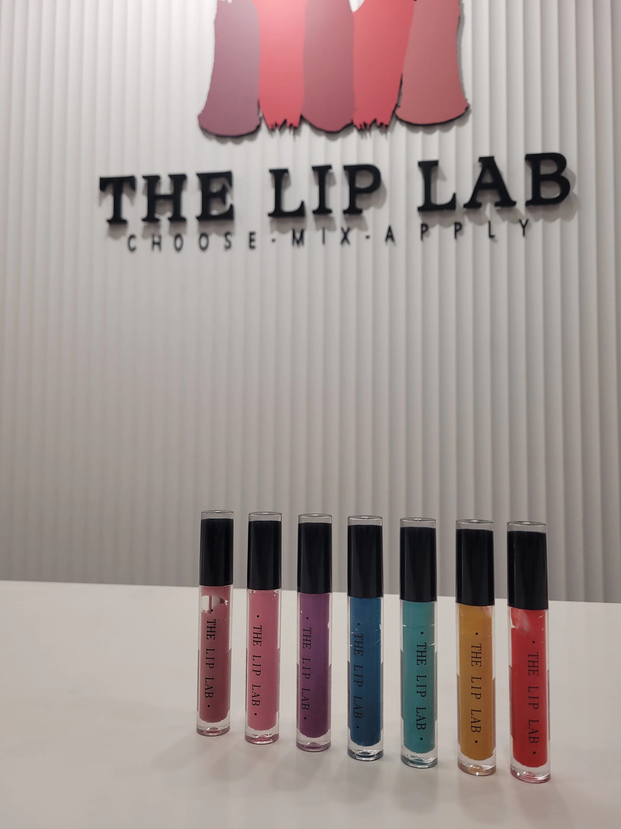 A row of lip gloss tubes in different pastel shades standing upright on a white surface, with a textured light-colored wall and signage in the background that reads 'THE LIP LAB - CHOOSE. MIX. APPLY'.
