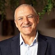A photo of Ray Galea speaker at the CCC Australia National Conference, smiling older man with gray hair and a dark suit, standing outdoors with greenery in the background.