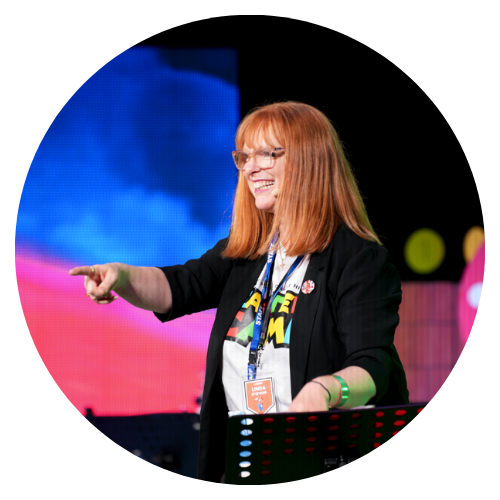Woman with red hair and glasses smiling and pointing on stage at a conference or event