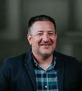 A smiling man with short hair and a beard, wearing a plaid shirt and a dark blazer, standing indoors against a neutral background.
