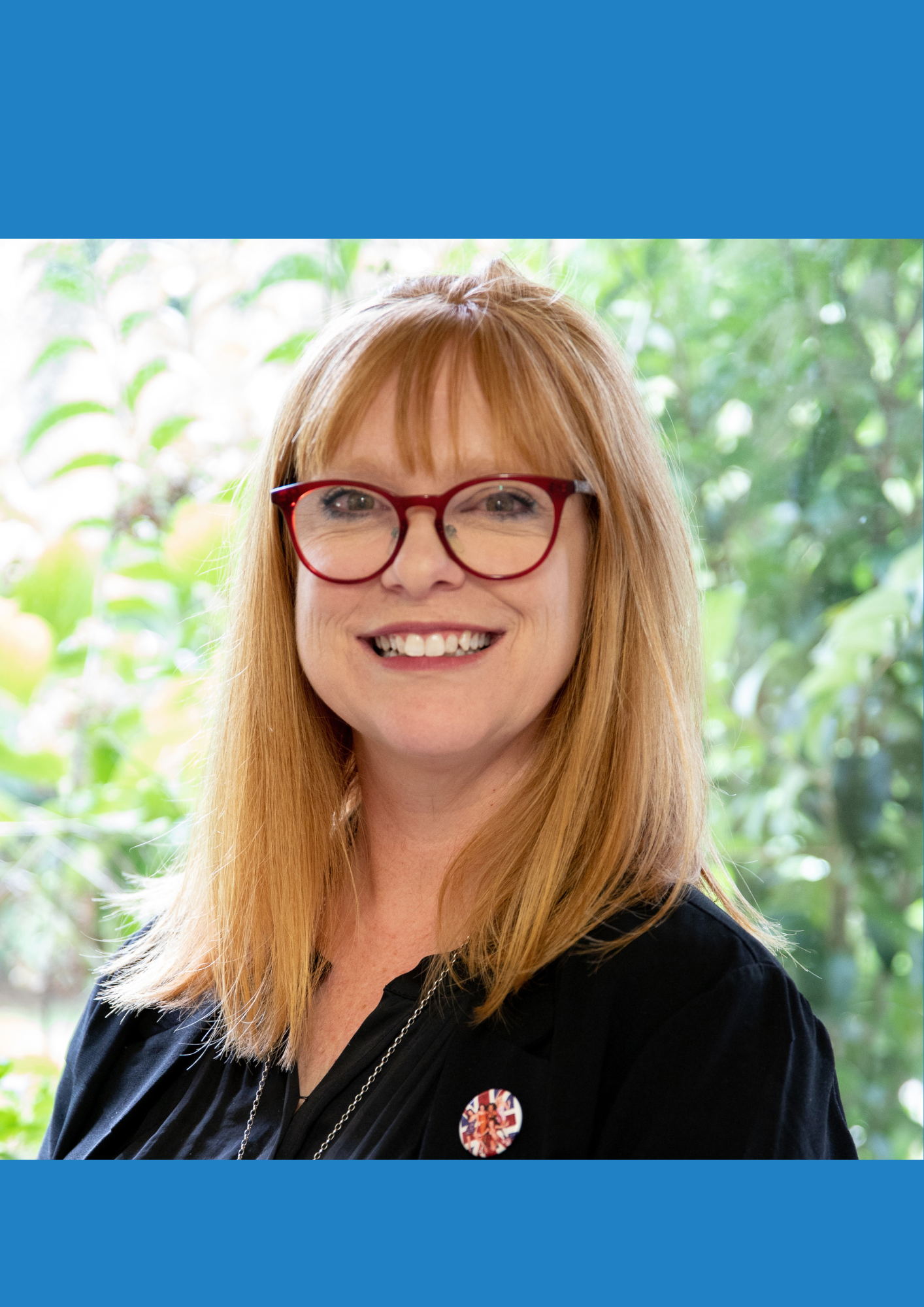 A photo of Linda Stephens a smiling woman with red hair, red glasses, wearing a black top with a Union Jack button, standing in front of greenery.