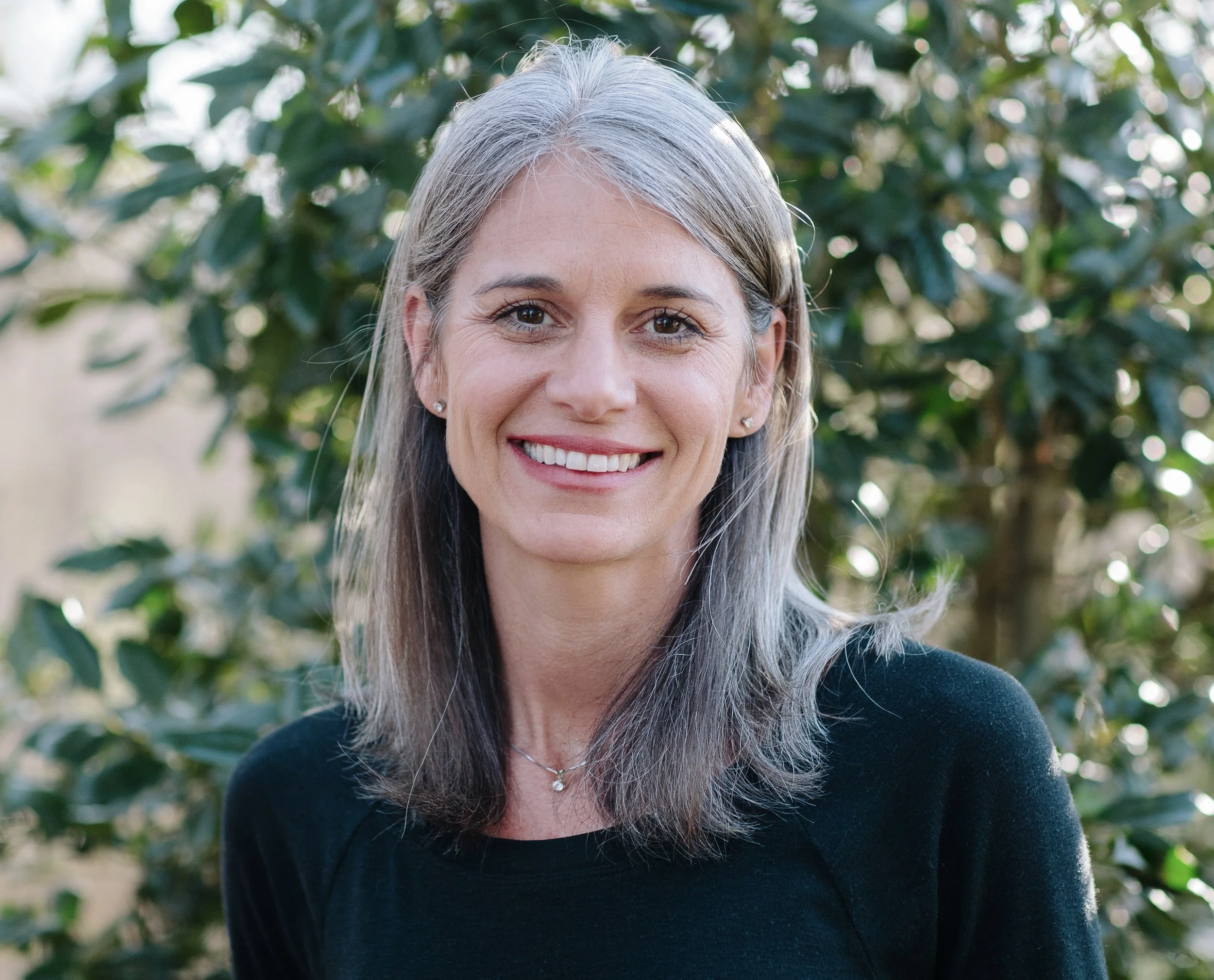 A woman with gray hair smiling outdoors in front of green leafy background.