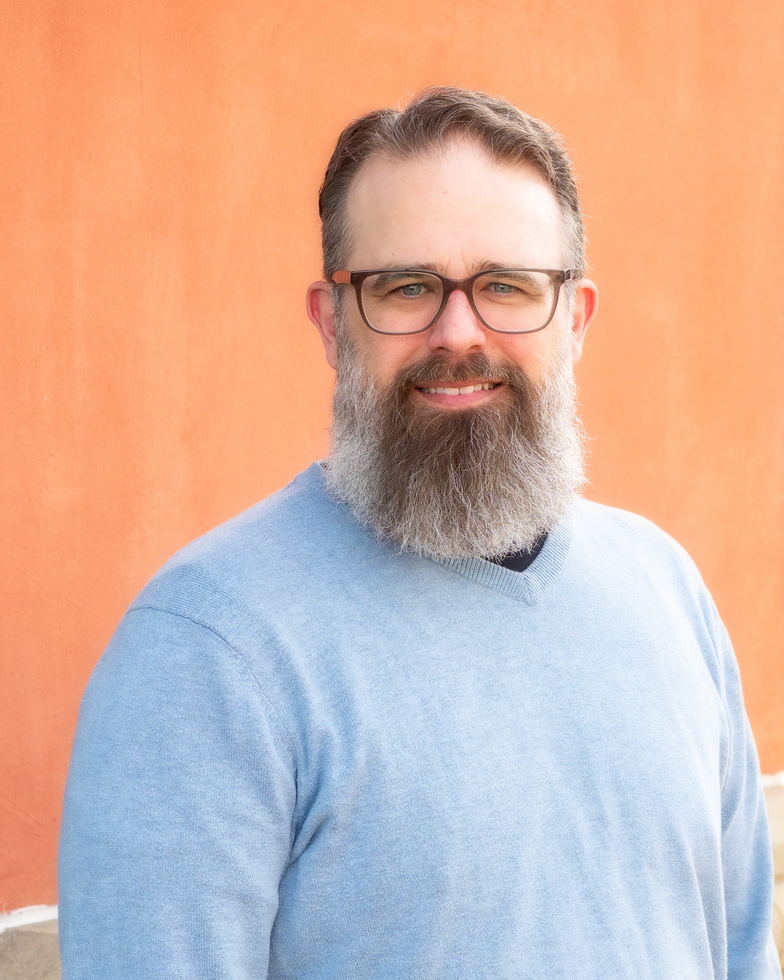Man with glasses and a beard wearing a light blue sweater, standing in front of an orange wall.