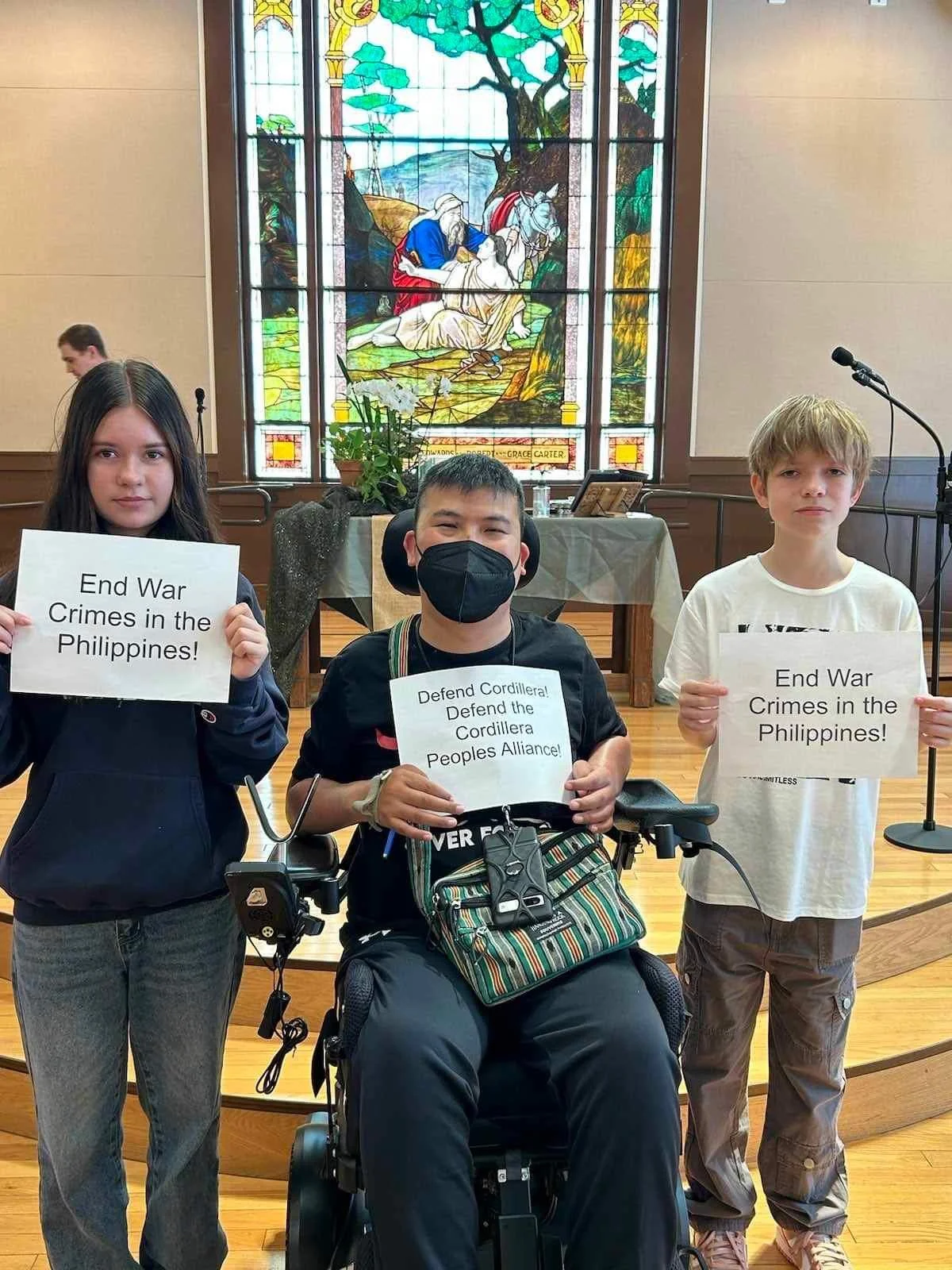 Brandon Lee holding a sign stating, “Defend Cordillera! Defend Cordillera Peoples Alliance!”. Two children on both sides of Brandon hold signs stating, “End War Crimes in the Philippines” during the 4th anniversary of his attempted assassination.