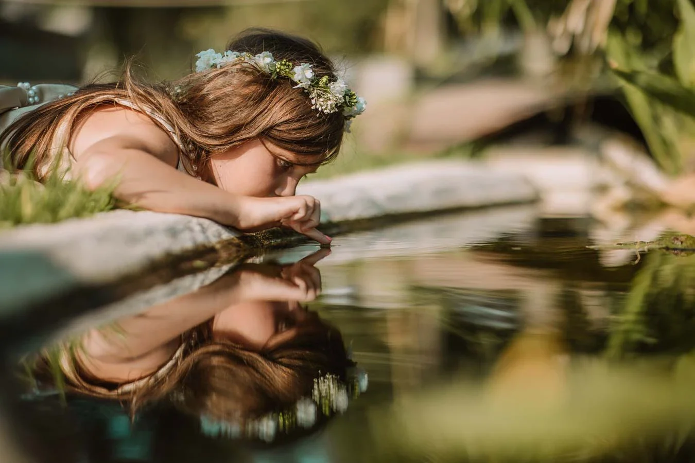 A young woman with a floral crown, lying on her stomach at the edge of a pond, drinking water from the pond with her finger.