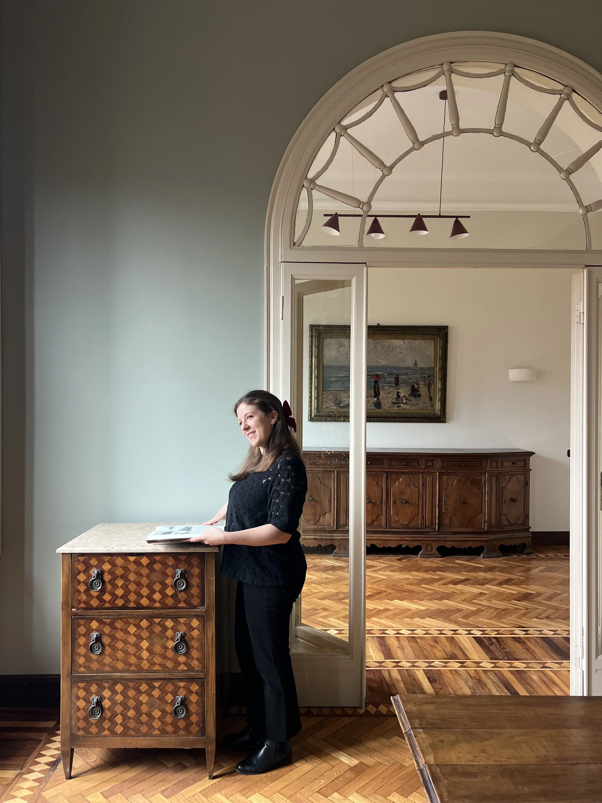 Una donna sorridente con capelli castani e vestita di nero, che si appoggia a un mobile antico con cassetti in legno a motivi geometrici, in una stanza elegante con pavimento in parquet e pareti chiare, all'interno di una casa o di un museo.