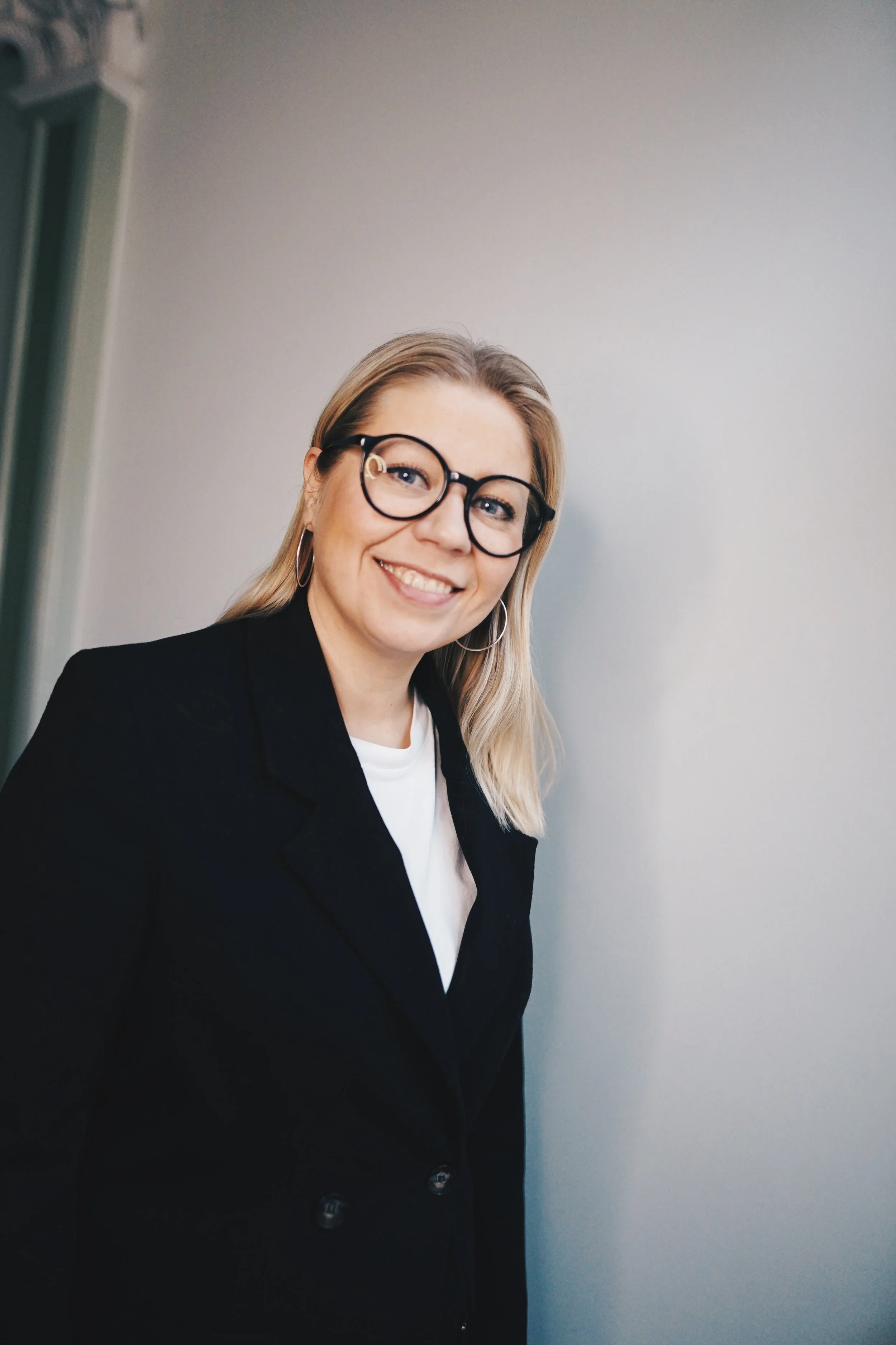 A woman with blonde hair, wearing glasses, hoop earrings, a black blazer, and a white top, smiling facing the camera.