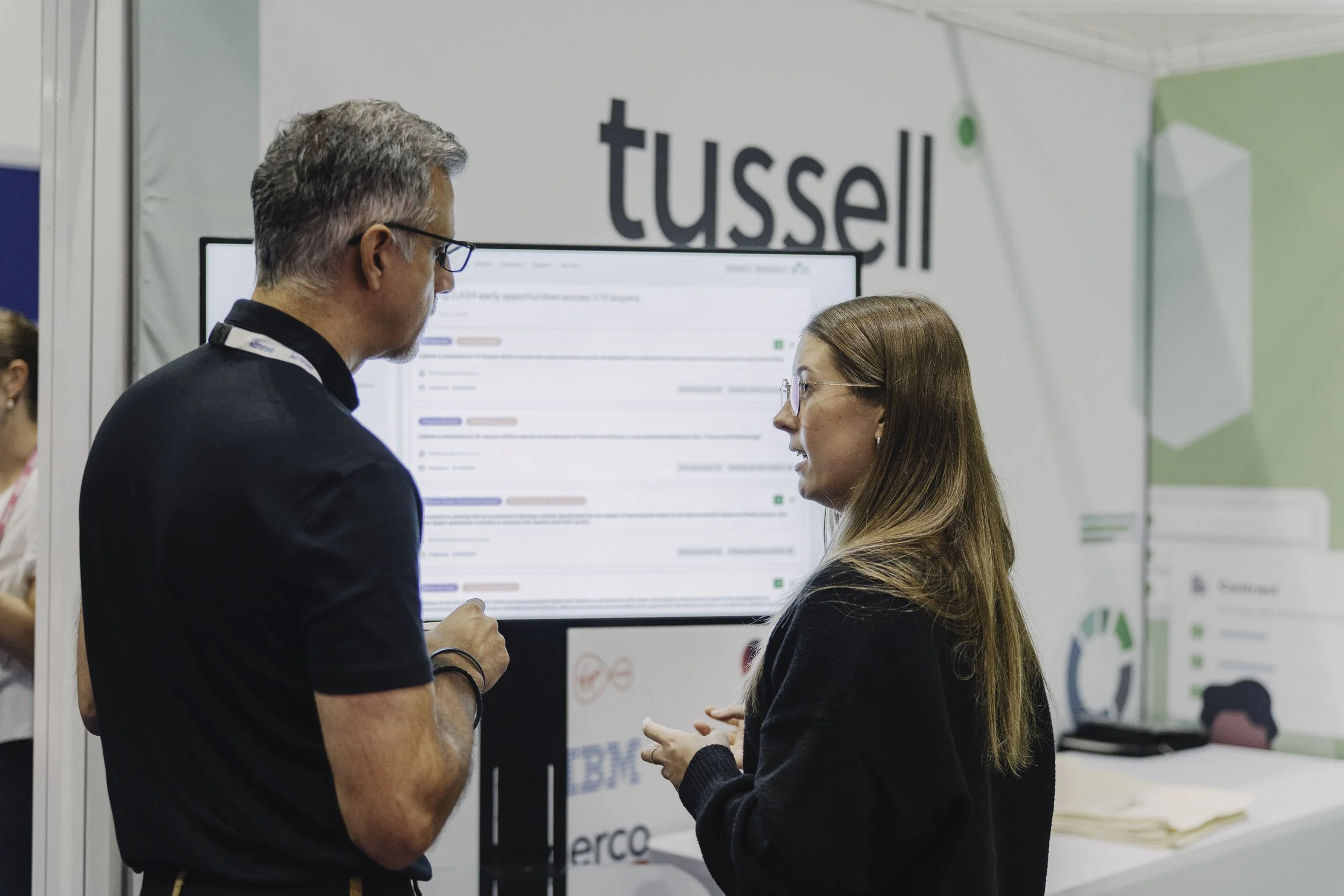 A man with gray hair and glasses and a woman with long brown hair and glasses engaging in conversation at a conference booth with the word 'tussell' on the wall behind them at DigiGov Expo.