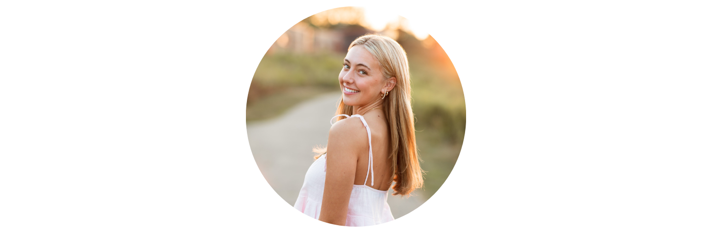 A person with long hair smiling and looking over their shoulder outdoors at sunset.