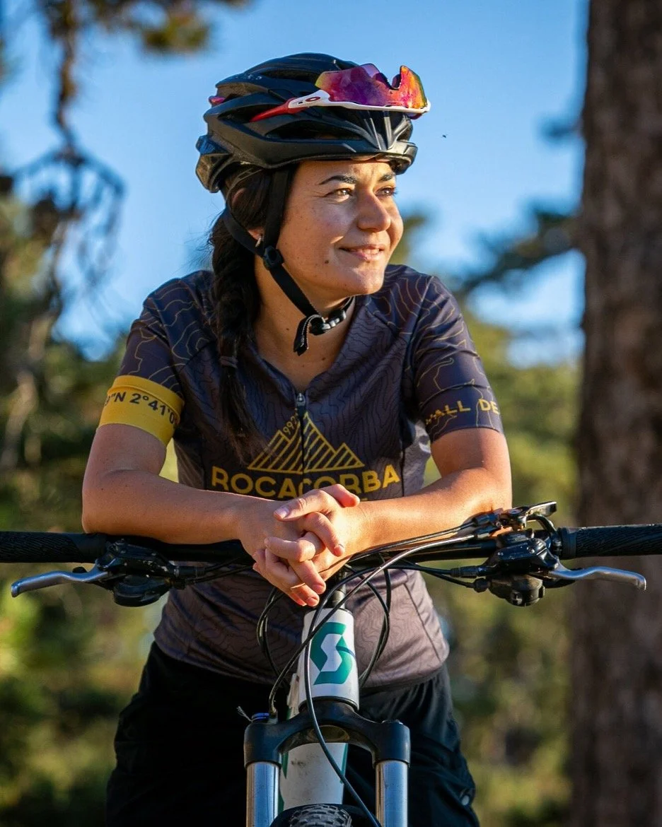 Woman wearing a black bike helmet with purple and orange sunglasses attached, standing with her mountain bike outdoors in a wooded area under a clear blue sky, smiling and relaxing.