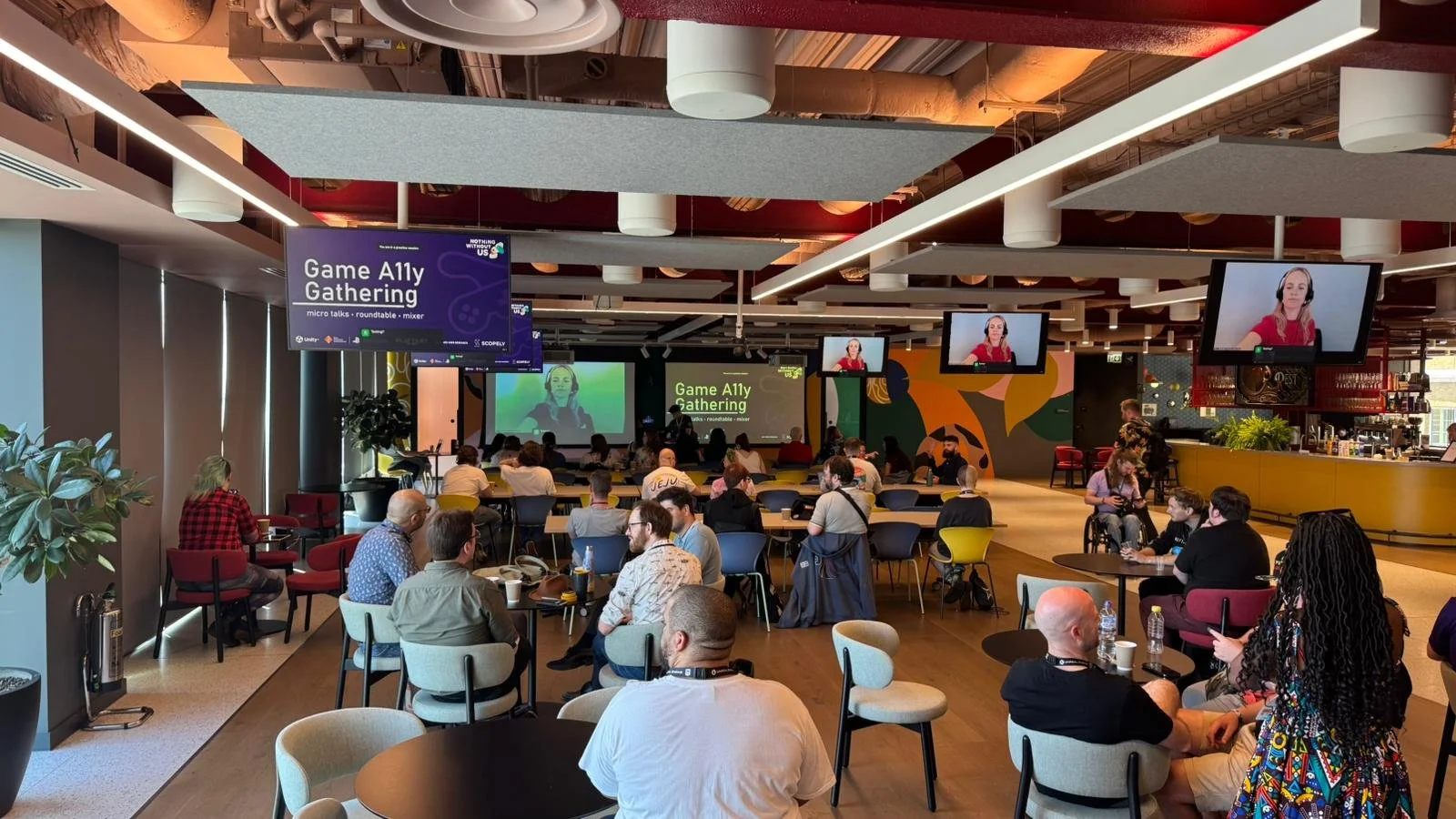A room with many tables and chairs, and a lot of attendees. Two large screens are at the back of the room, one shows the talk slides and one shows a BSL interpreter.