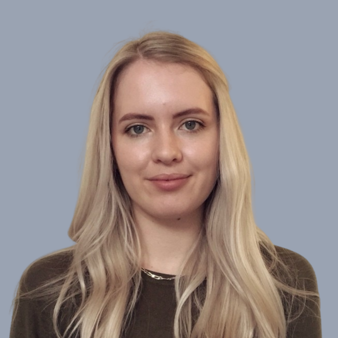 Portrait of a young woman with long blonde hair, blue eyes, and light makeup, wearing a black top and a silver chain necklace, against a plain gray background.