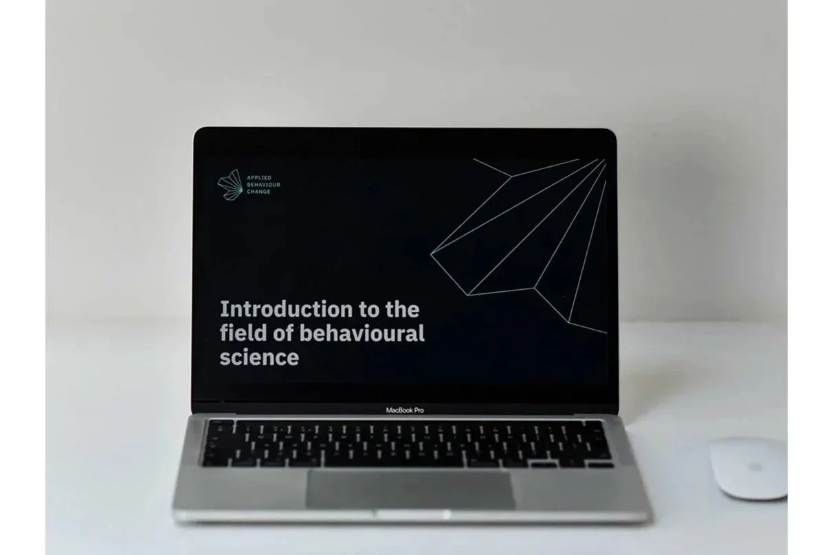 Laptop displaying a presentation slide titled "Introduction to the field of behavioural science" with a logo in the top left corner and a geometric graphic in the top right corner, on a white surface with a computer mouse nearby.