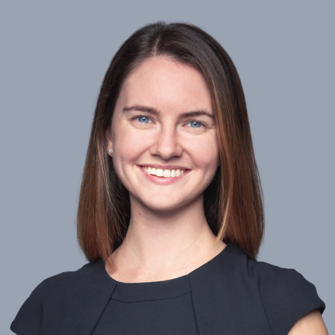 A headshot of a smiling woman with shoulder-length brown hair, blue eyes, wearing a black top, and earrings, against a plain gray background.